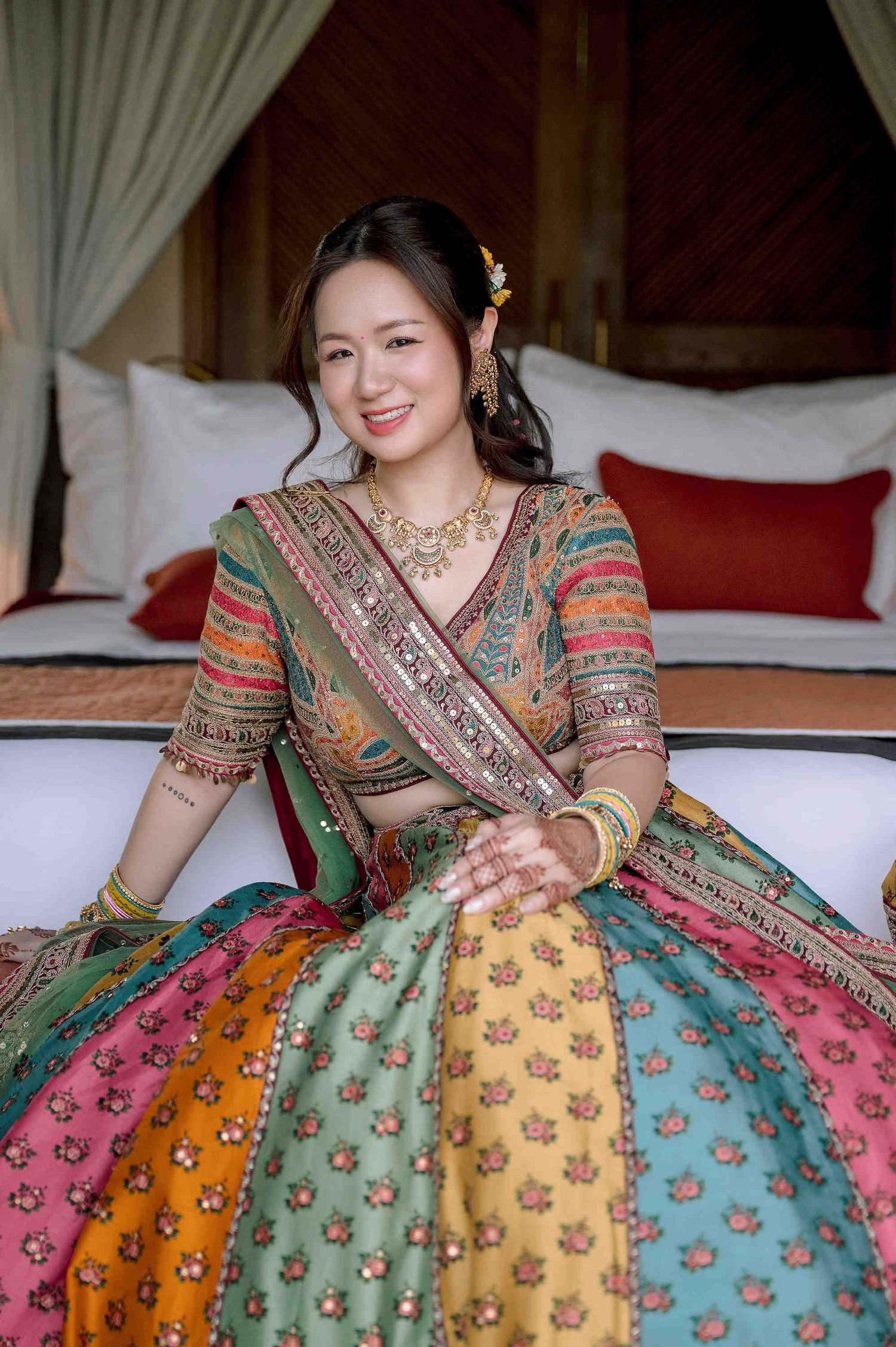 Bride in colorful multi-panel lehenga with gold jewelry smiles while seated at an Indian wedding celebration.