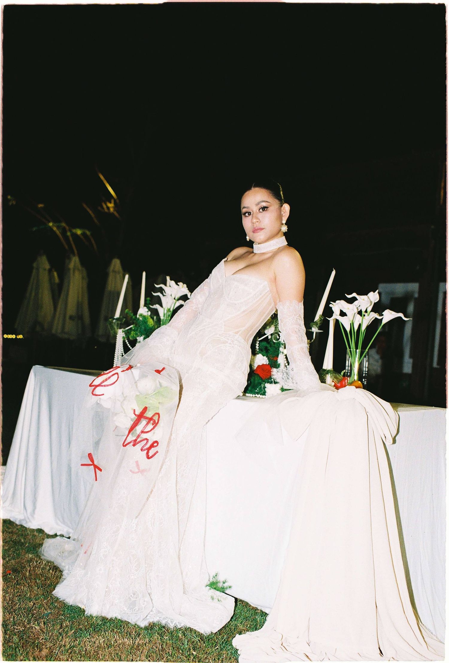 Saigon pre-wedding: elegant bridal portrait beside a white-clothed table with floral arrangements in an evening garden, by AnhVuWedding.