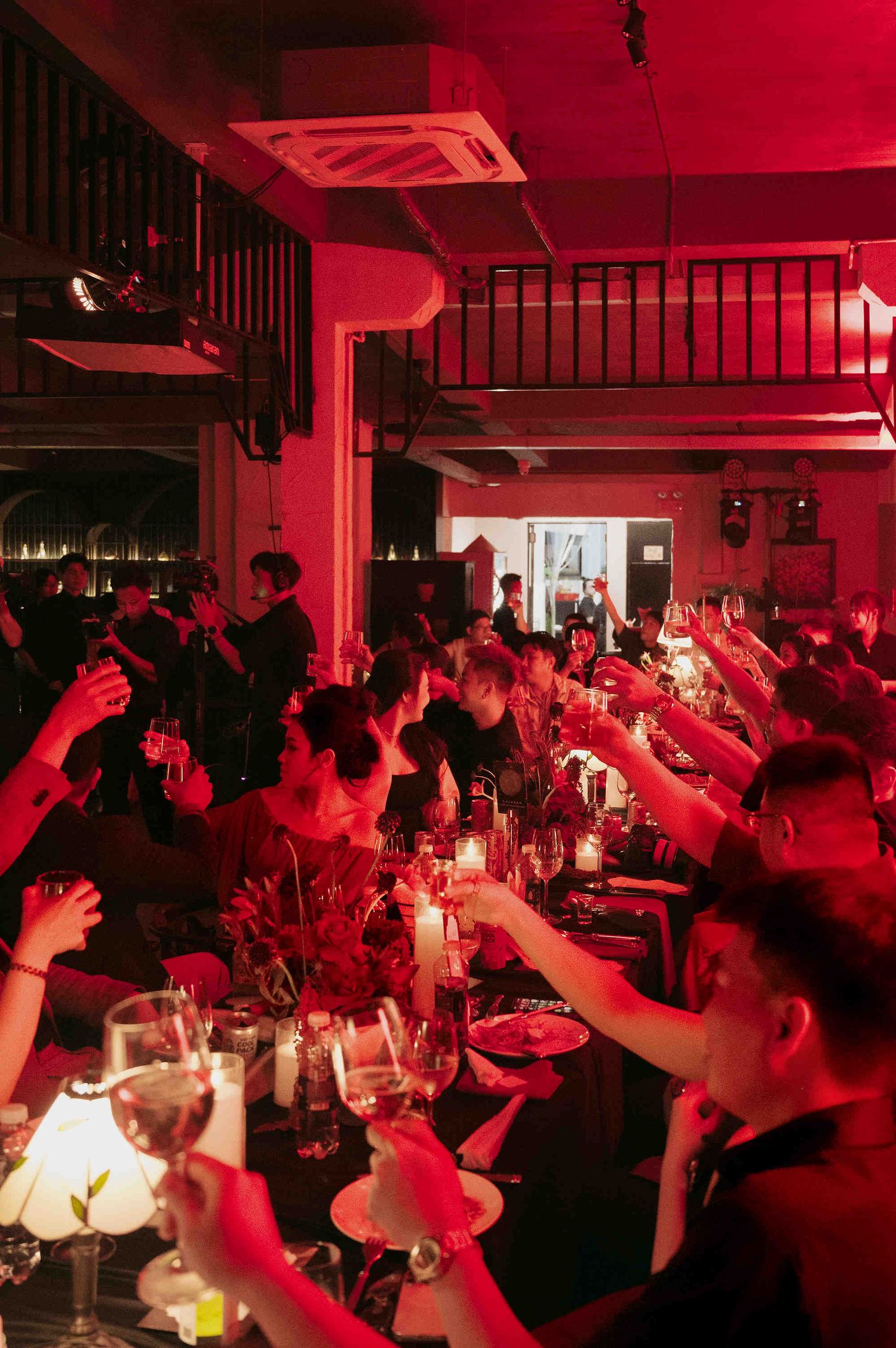 Guests raise glasses in a toast at a dimly lit dinner party bathed in dramatic red lighting.