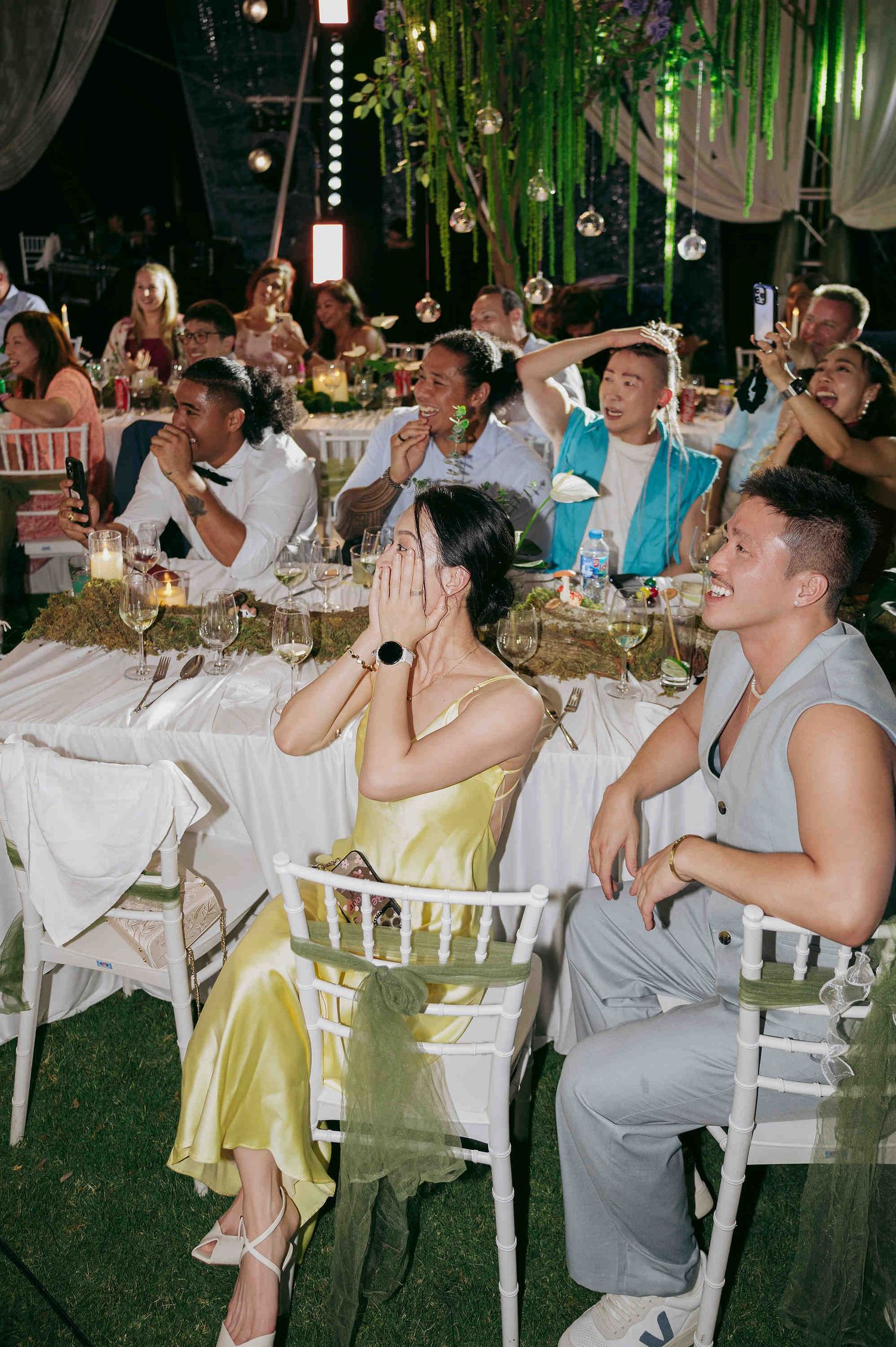 Guests laughing and reacting emotionally at an elegant outdoor wedding reception with floral decorations.