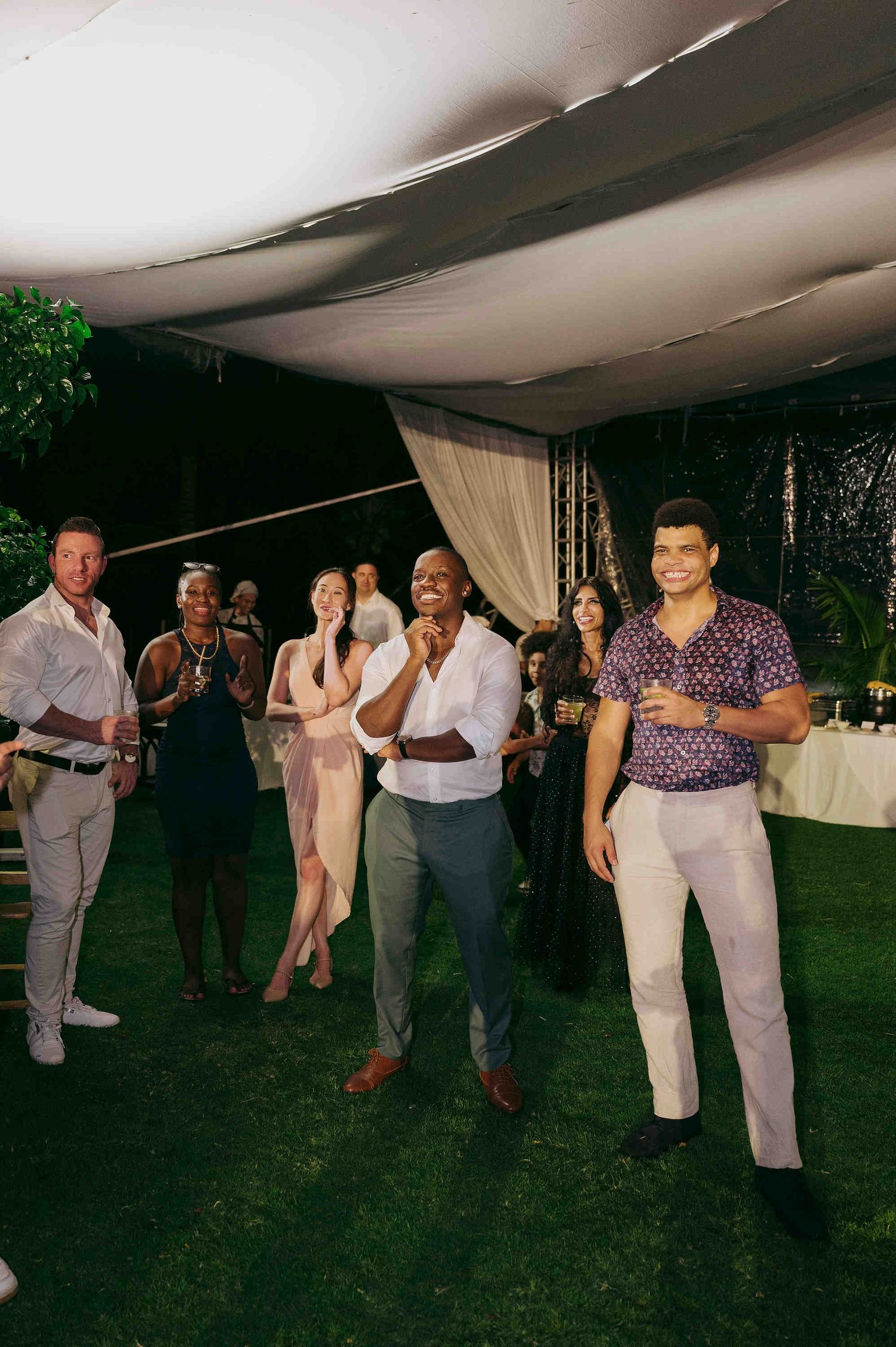 Guests laughing and dancing at an outdoor evening wedding reception under a white tent on green grass.