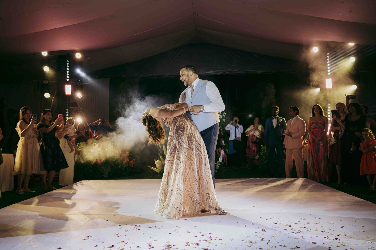 Bride and groom share first dance on white dance floor surrounded by guests at elegant outdoor wedding reception.