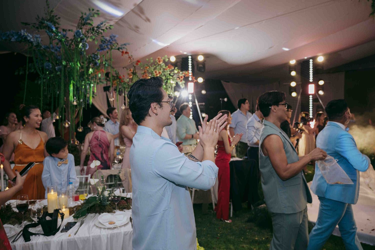 Guests clapping and celebrating at an elegant outdoor wedding reception with string lights and floral decorations.