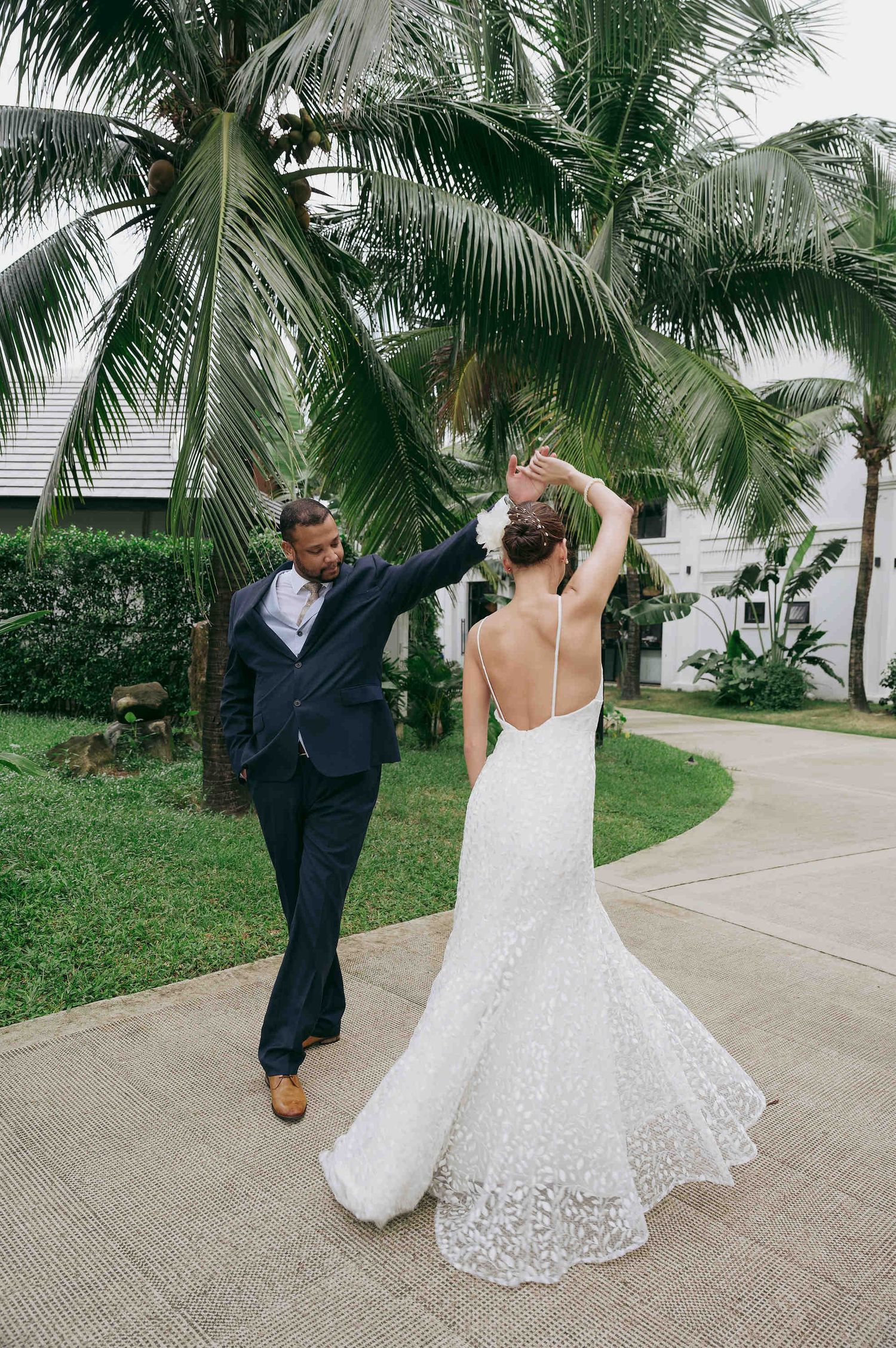 Bride in lace gown spins as groom in navy suit twirls her under palm trees on tropical wedding day.