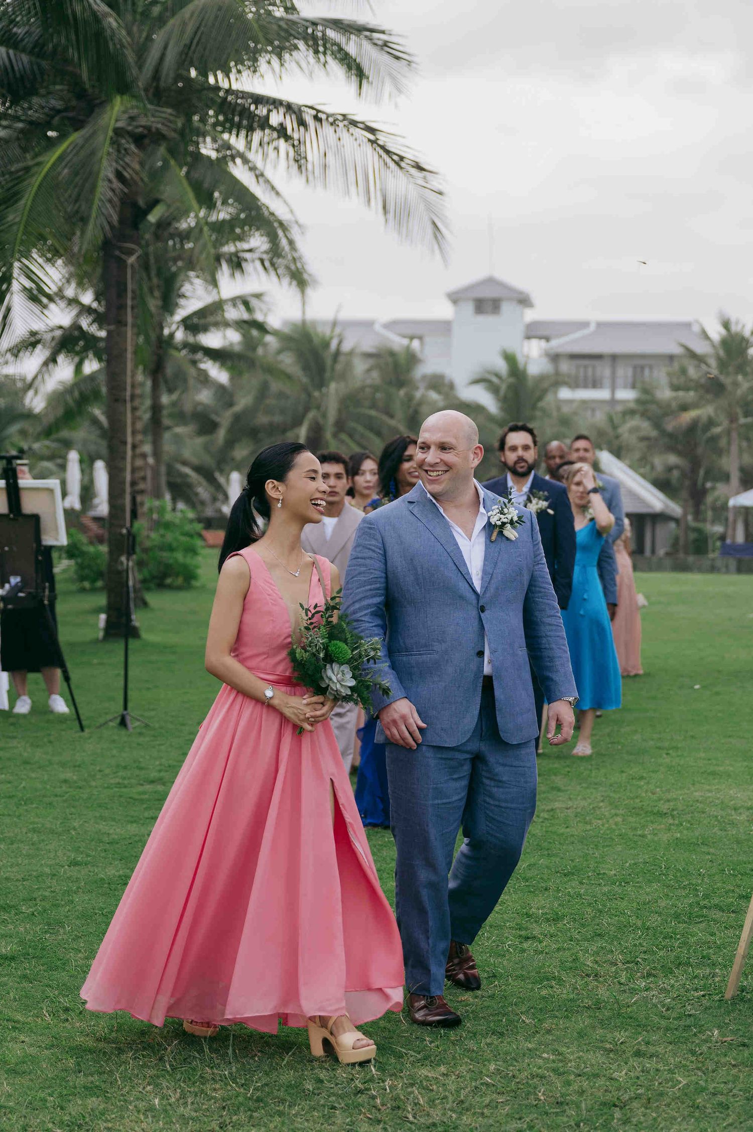 Bride in pink dress and groom in blue suit walk down outdoor aisle at tropical beach wedding ceremony.