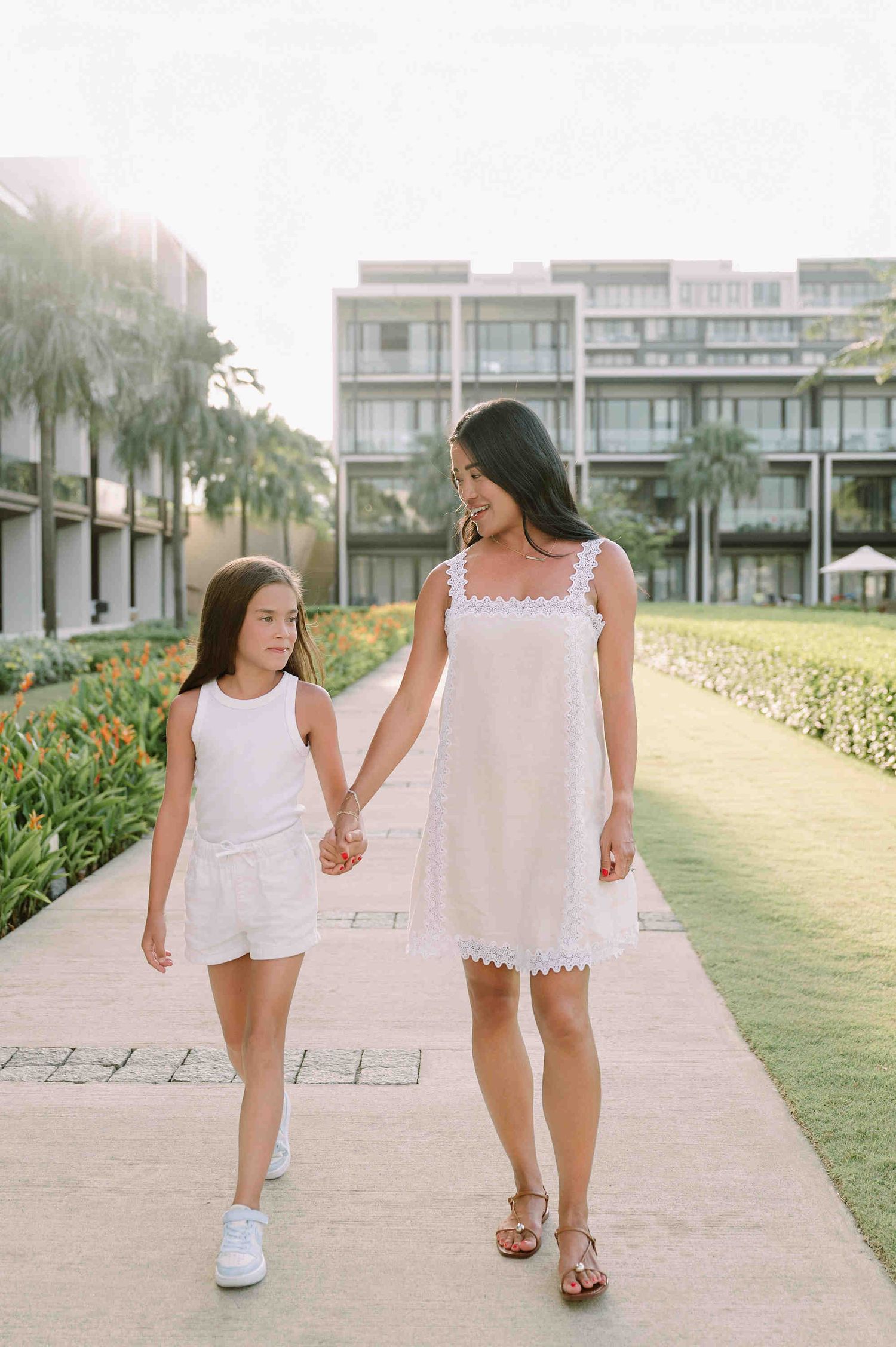 Da Nang family: mother and daughter walking hand in hand along a beachside path in matching white summer outfits, by AnhVuWedding.
