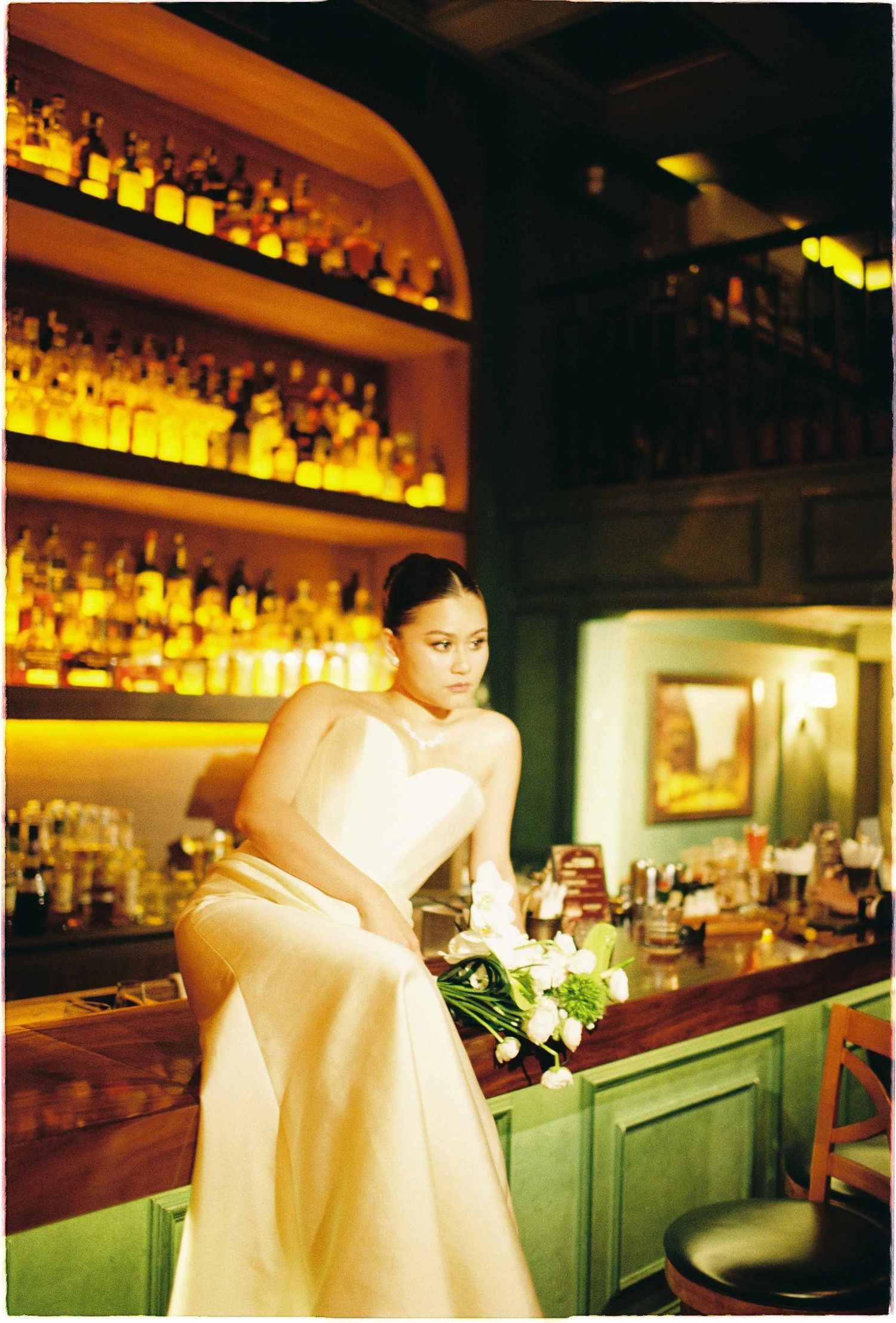 Vietnam pre-wedding: bride in white dress poses elegantly at bar with illuminated bottle display shelves, by AnhVuWedding.