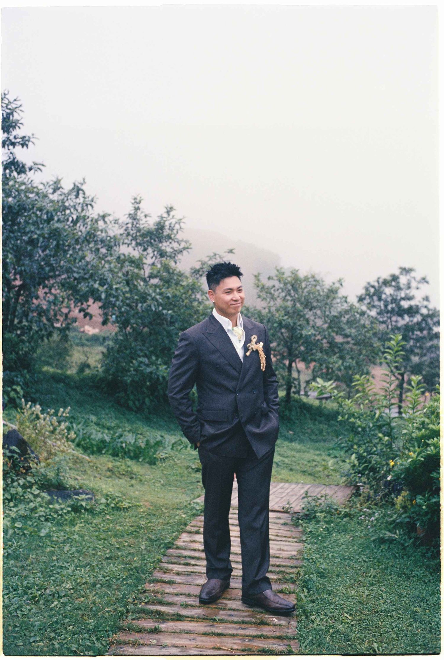wedding: person in a black suit stands on a wooden walkway surrounded by lush greenery and flowers, by AnhVuWedding.