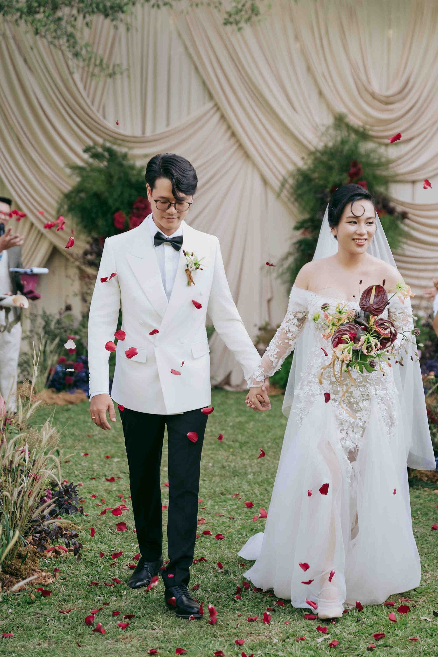 Bride and groom walk down aisle holding hands, rose petals falling, white tuxedo and lace wedding gown, outdoor ceremony.