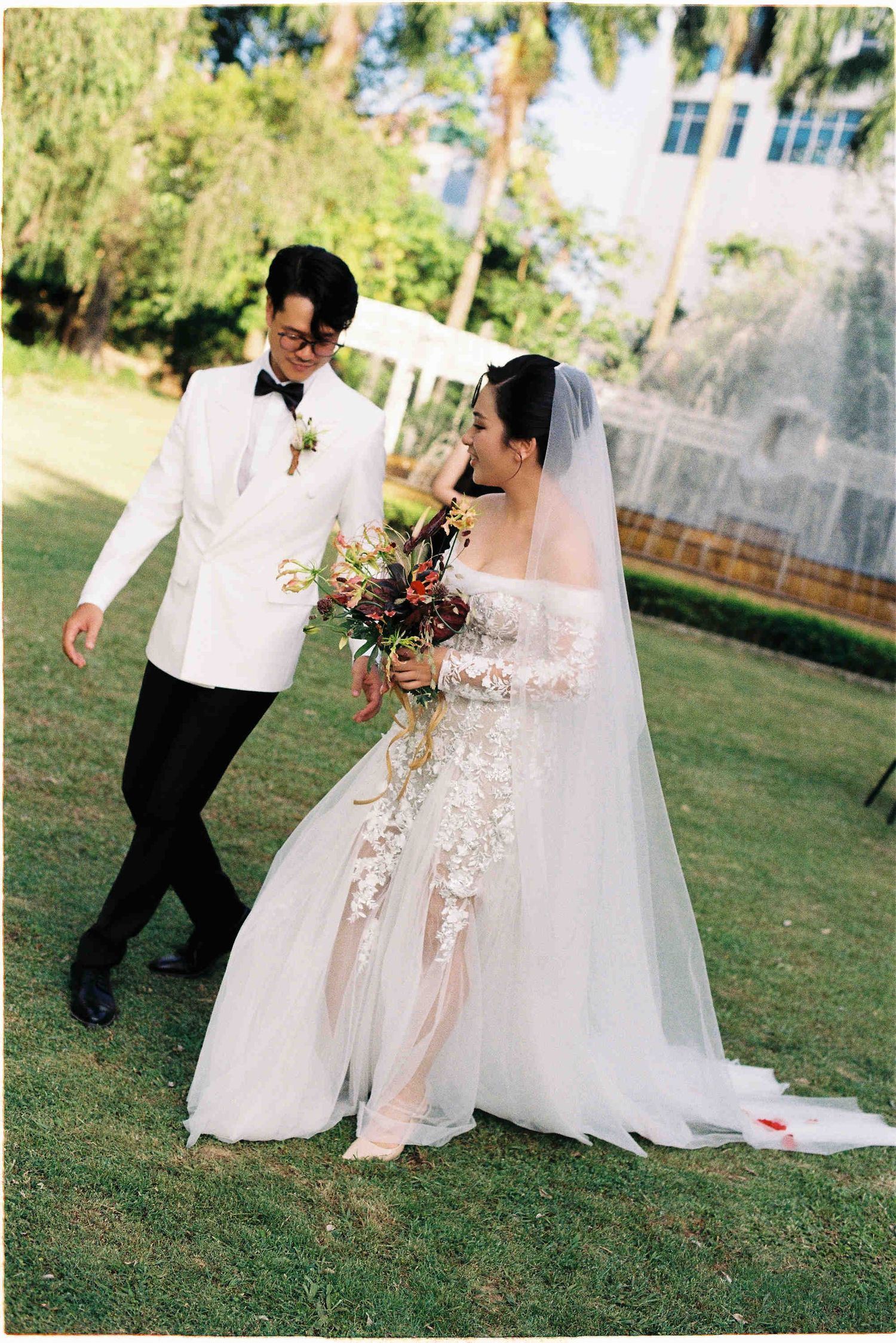 Bride in lace wedding gown with veil holds bouquet beside groom in white tuxedo on green lawn.