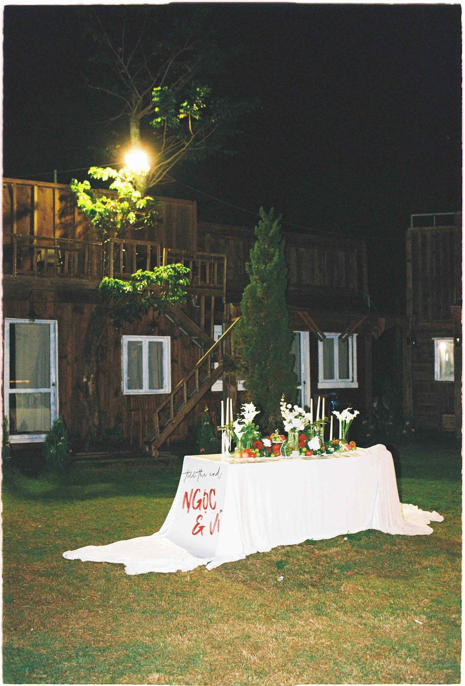 Saigon pre-wedding: outdoor nighttime scene showing white-clothed tables set up on a dark lawn near buildings, by AnhVuWedding.