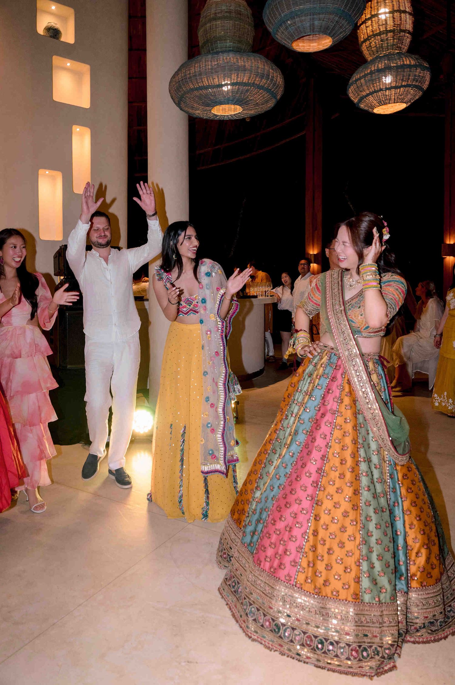 Guests in vibrant Indian attire dance joyfully at a wedding celebration with ornate hanging lanterns.