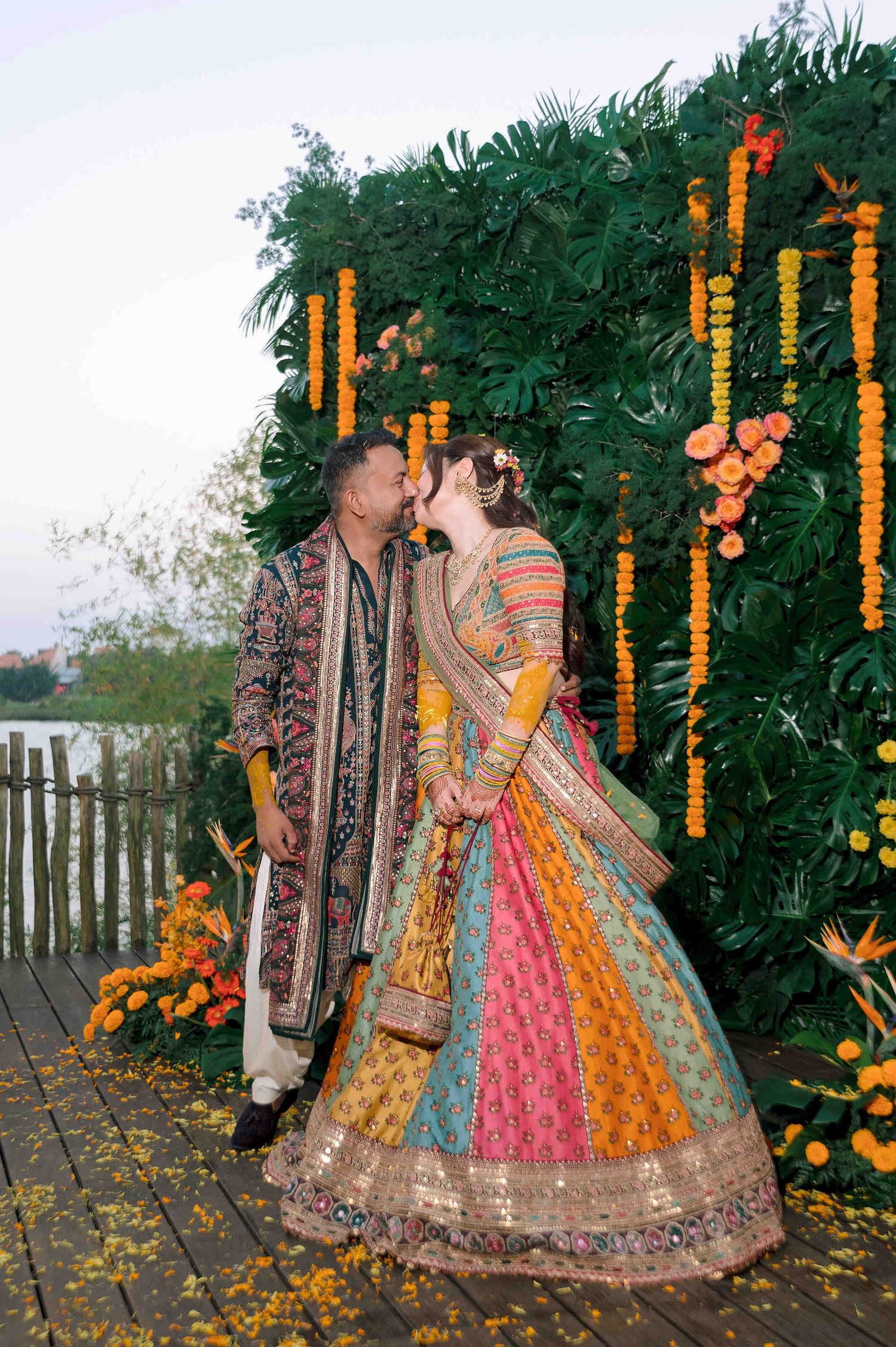 Couple sharing a kiss in traditional Indian wedding attire before a floral marigold backdrop.