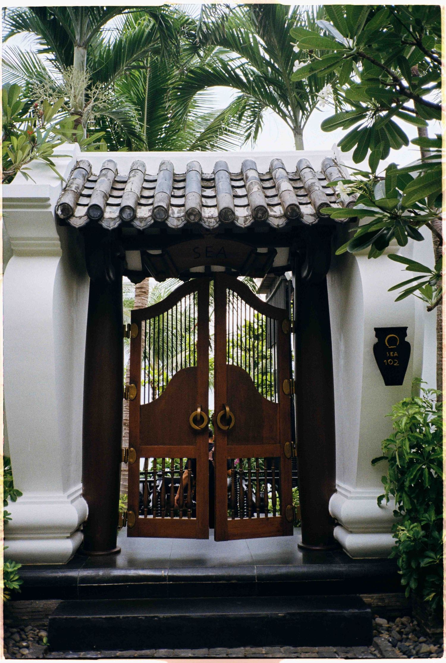 Da Nang photo: ornate wooden double gates with Asian-style tiled roof entrance flanked by white columns and lush tropical, by AnhVuWedding.
