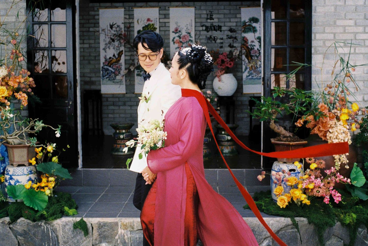 Couple in traditional Vietnamese ao dai attire pose outside a decorated venue holding a bridal bouquet.