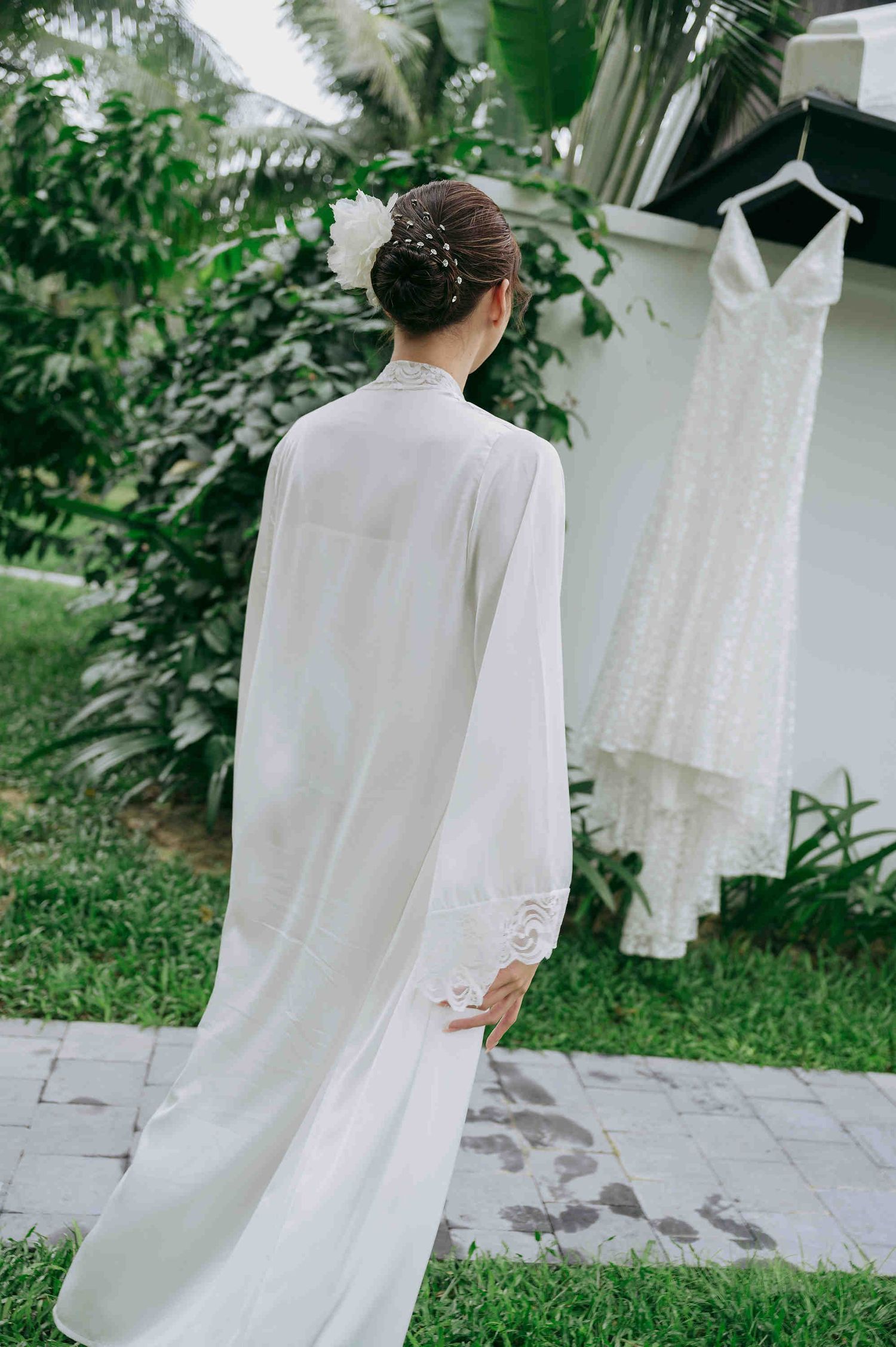 Bride in white robe views her lace wedding dress hanging outdoors among lush green tropical foliage.