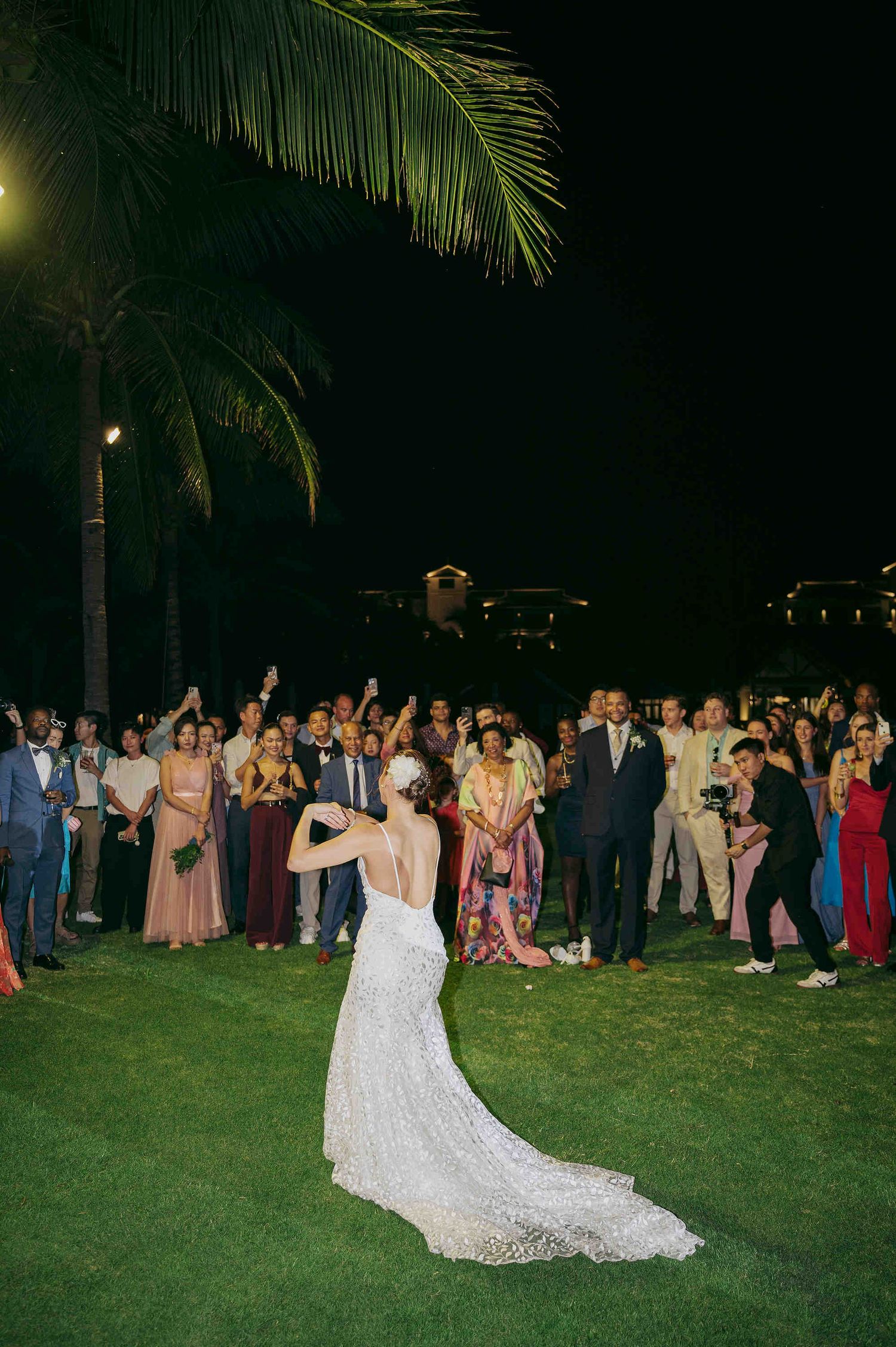 Bride in lace gown tosses bouquet to excited wedding guests on tropical lawn at night.