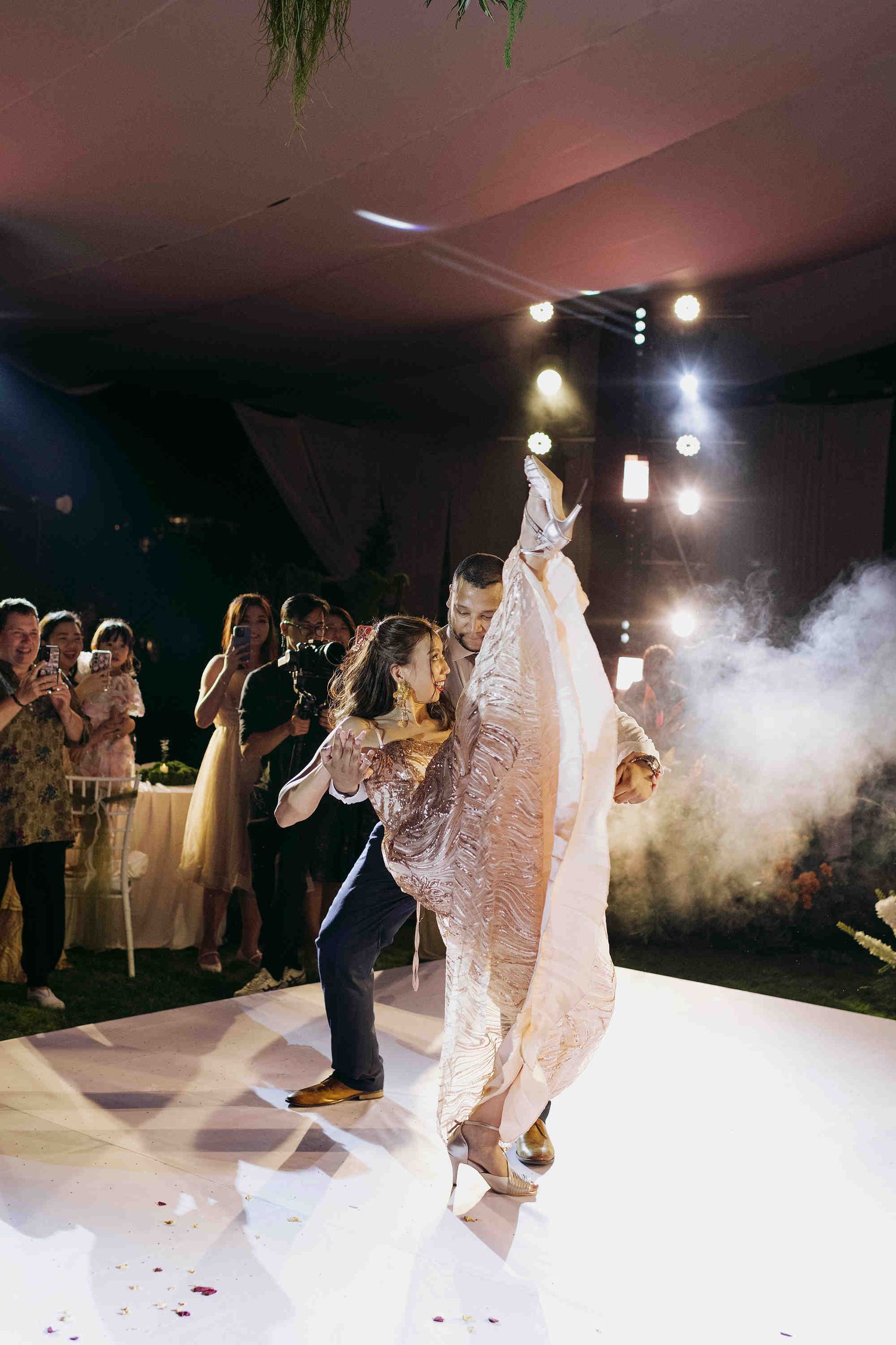 Bride and groom share first dance on white dance floor with smoke effects as wedding guests watch and photograph the moment.