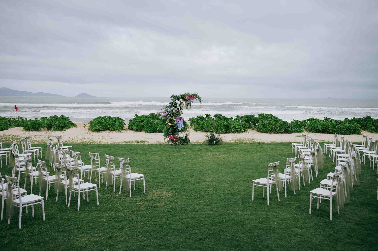 Beach wedding ceremony setup with white chairs on green lawn, floral arch, and ocean backdrop under overcast skies.