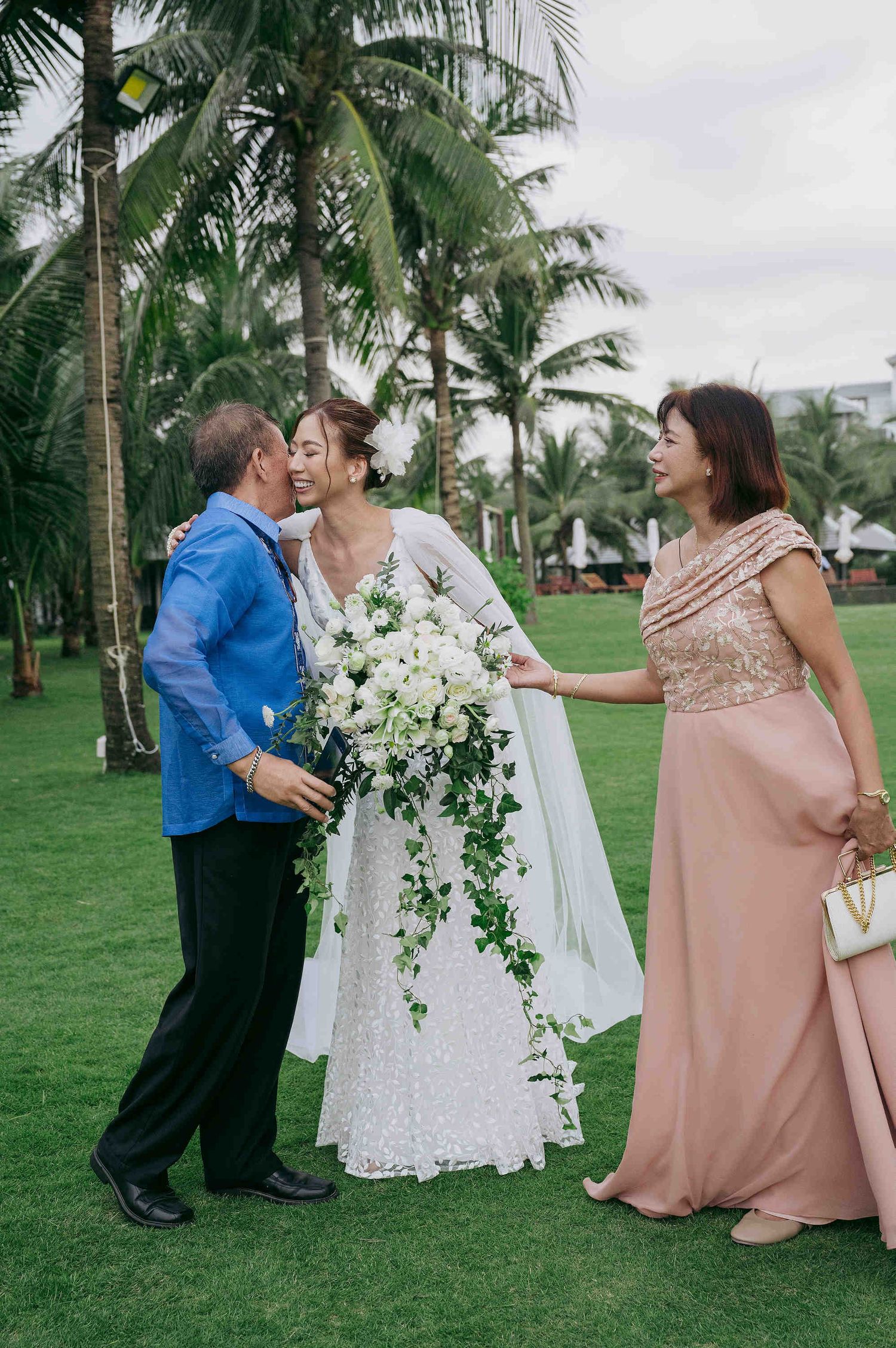 Bride in white lace gown holding cascading bouquet receives kiss from father while mother in pink gown smiles outdoors.