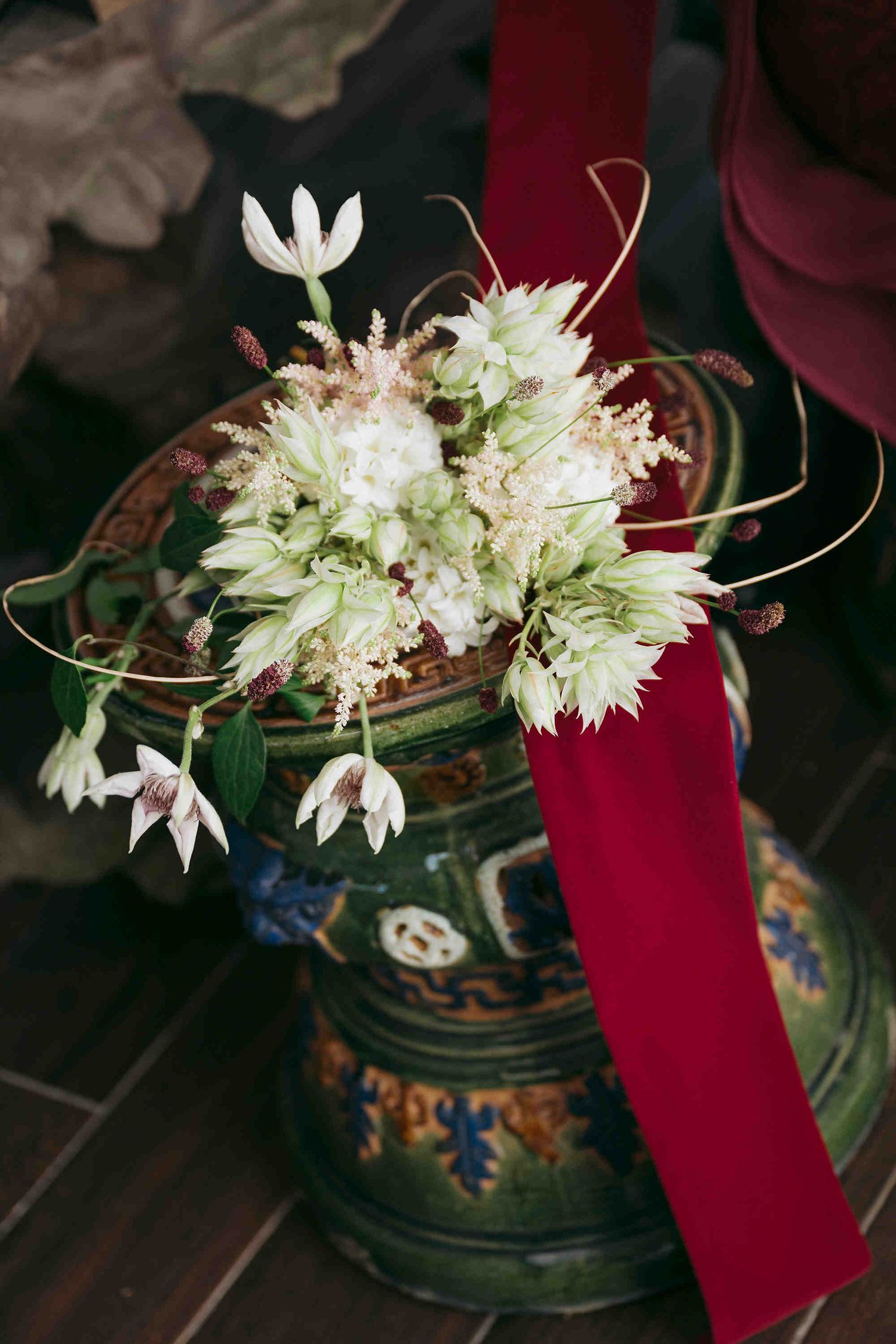 Ornate ceramic vase with white wildflowers and green hops, accented by a deep red velvet ribbon on a wooden surface.