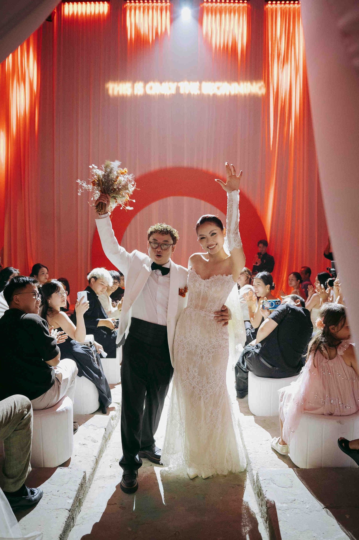 Joyful newlyweds celebrate at a modern wedding ceremony with red drapes and neon sign backdrop.