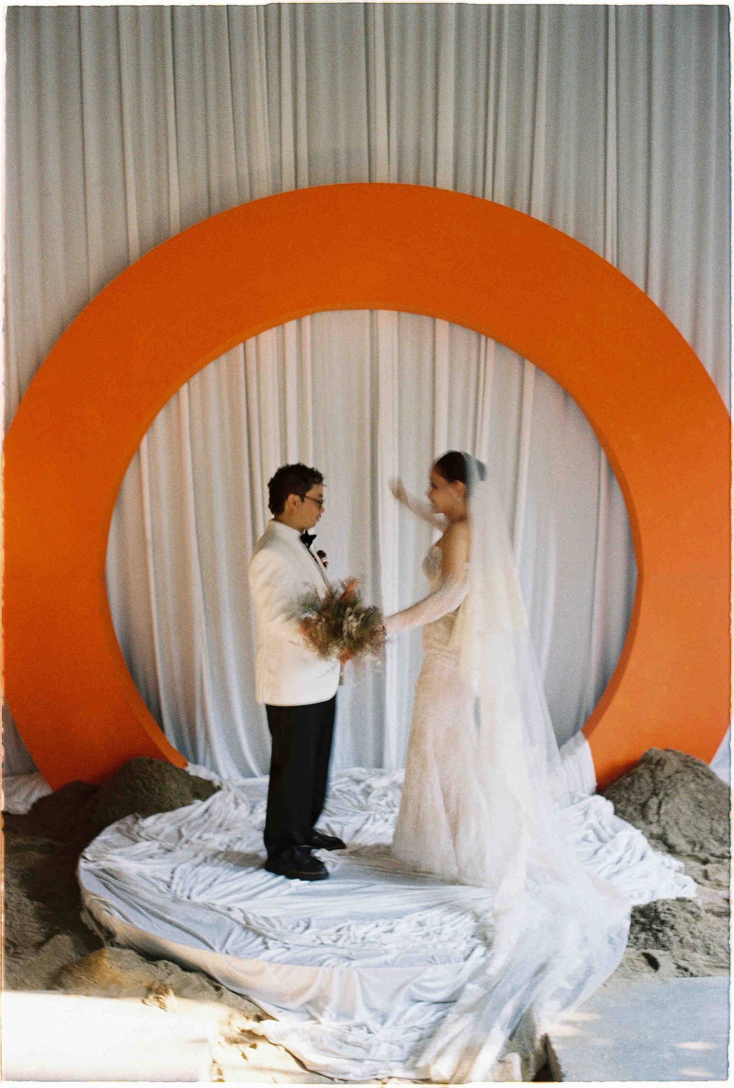 Bride and groom exchange vows beneath a bold orange circular arch with white drape backdrop at an outdoor wedding.