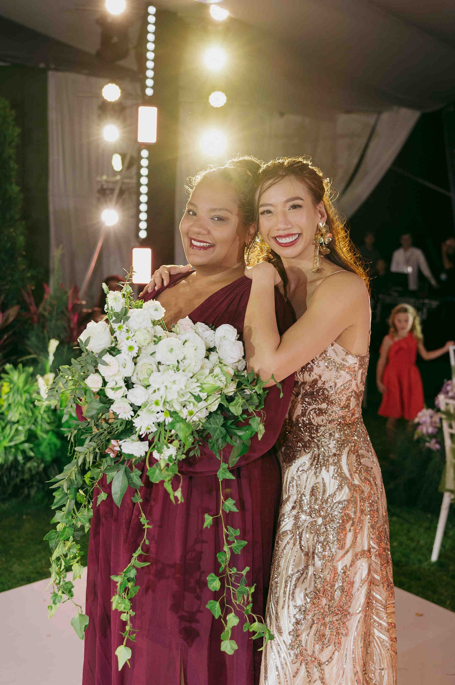 Two women in elegant gowns smile at a wedding reception, one holding a lush white floral bouquet with trailing greenery.