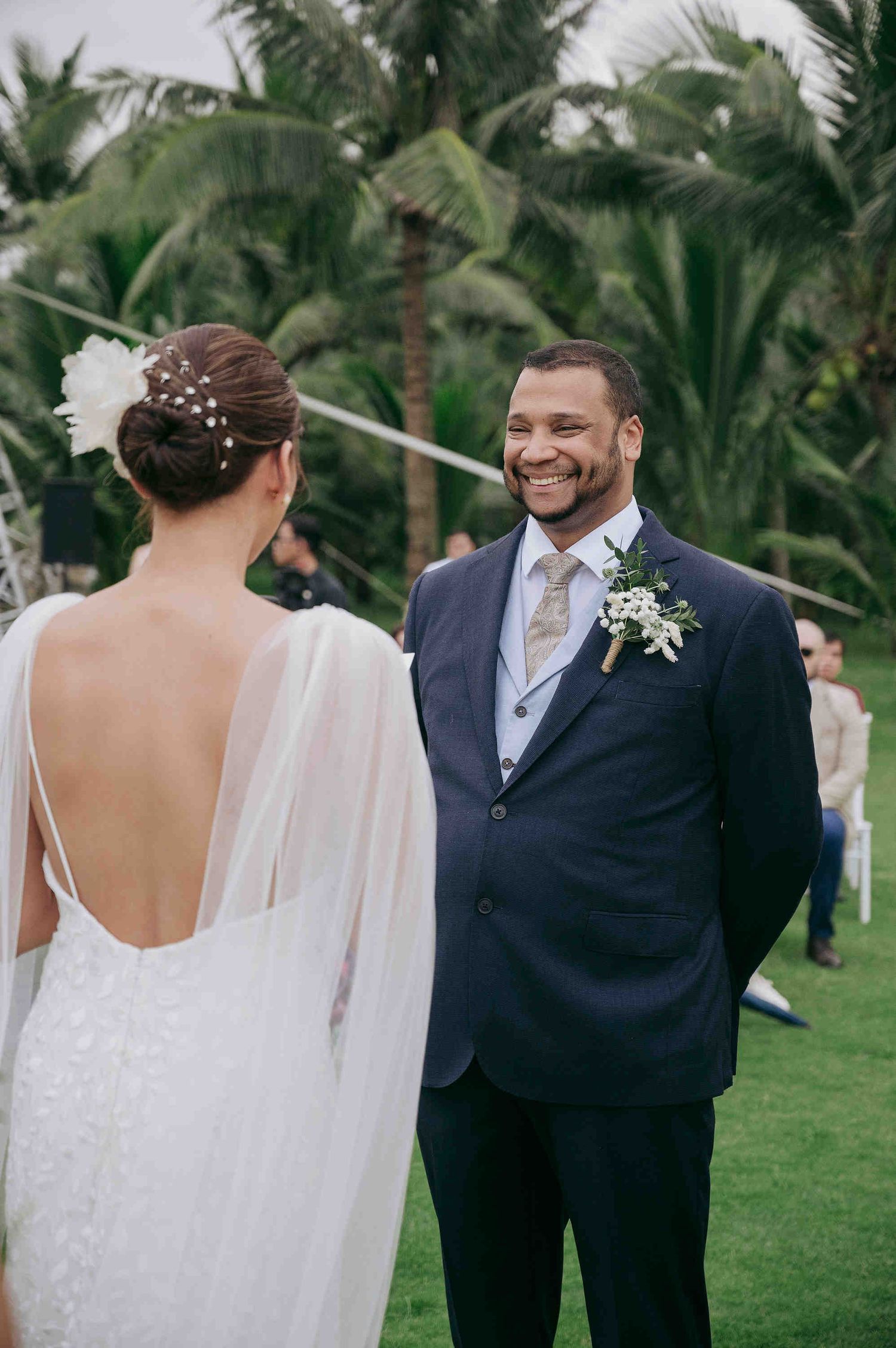 Groom in navy suit smiling warmly at bride in backless lace gown at tropical outdoor wedding.