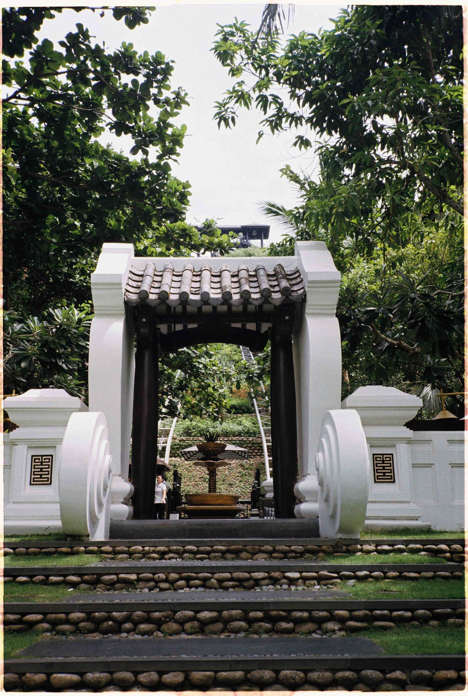 Da Nang photo: white ornate Asian garden gate with dark tiled roof arch surrounded by lush tropical trees and stone, by AnhVuWedding.