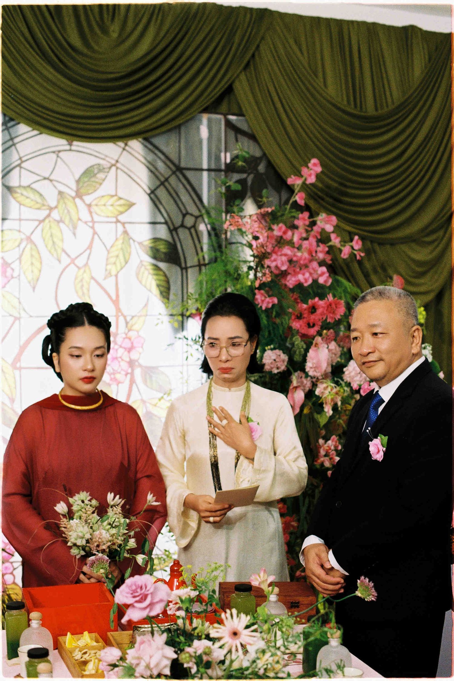 Three people stand by a floral table at an Asian ceremony, woman in red ao dai, man in suit, decorated backdrop.