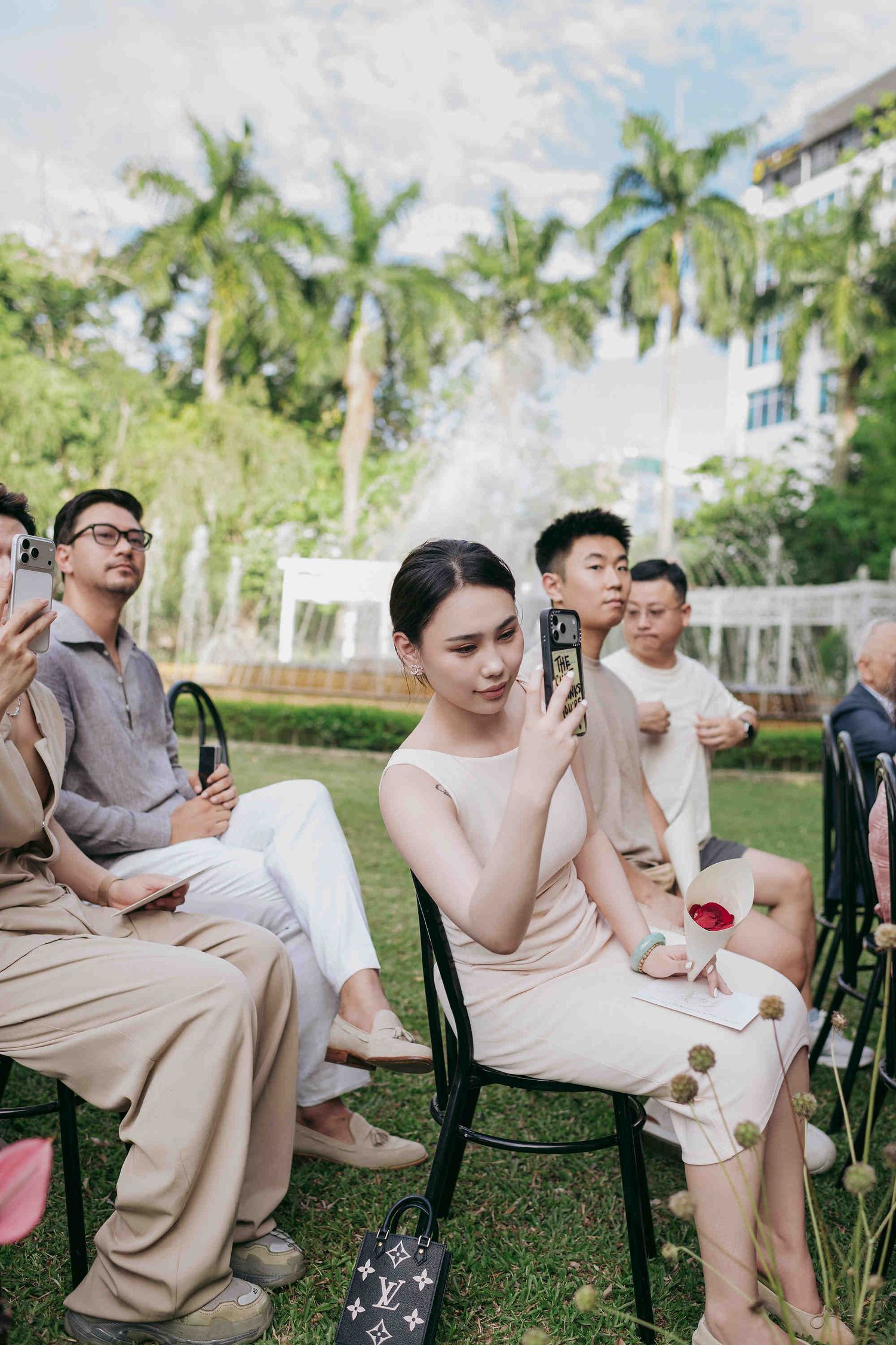 Guests seated outdoors at a garden wedding ceremony, with a woman in beige taking photos on her phone.