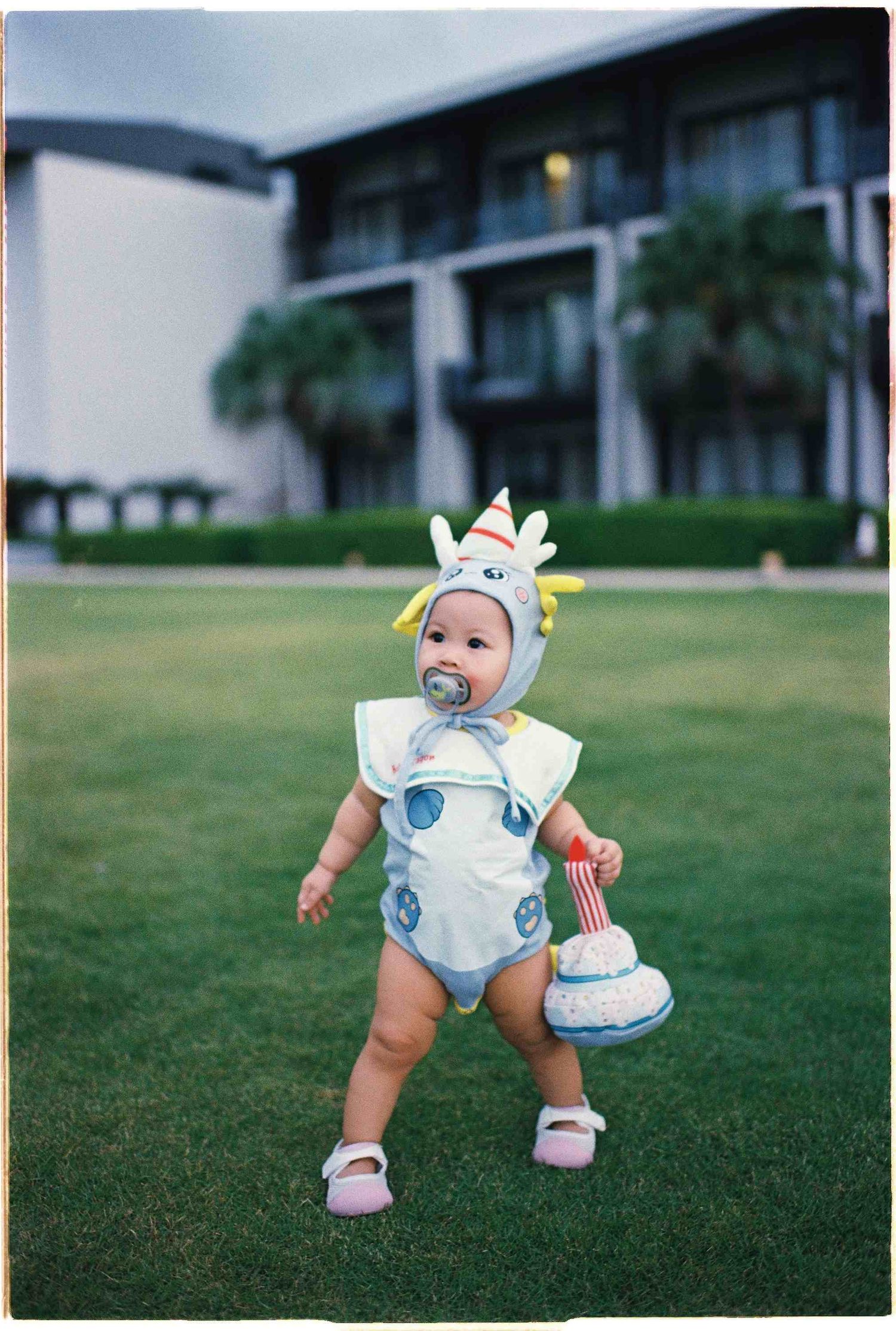 Da Nang family: baby in an adorable animal costume with a matching hat toddles on grass with a resort building backdrop, by AnhVuWedding.