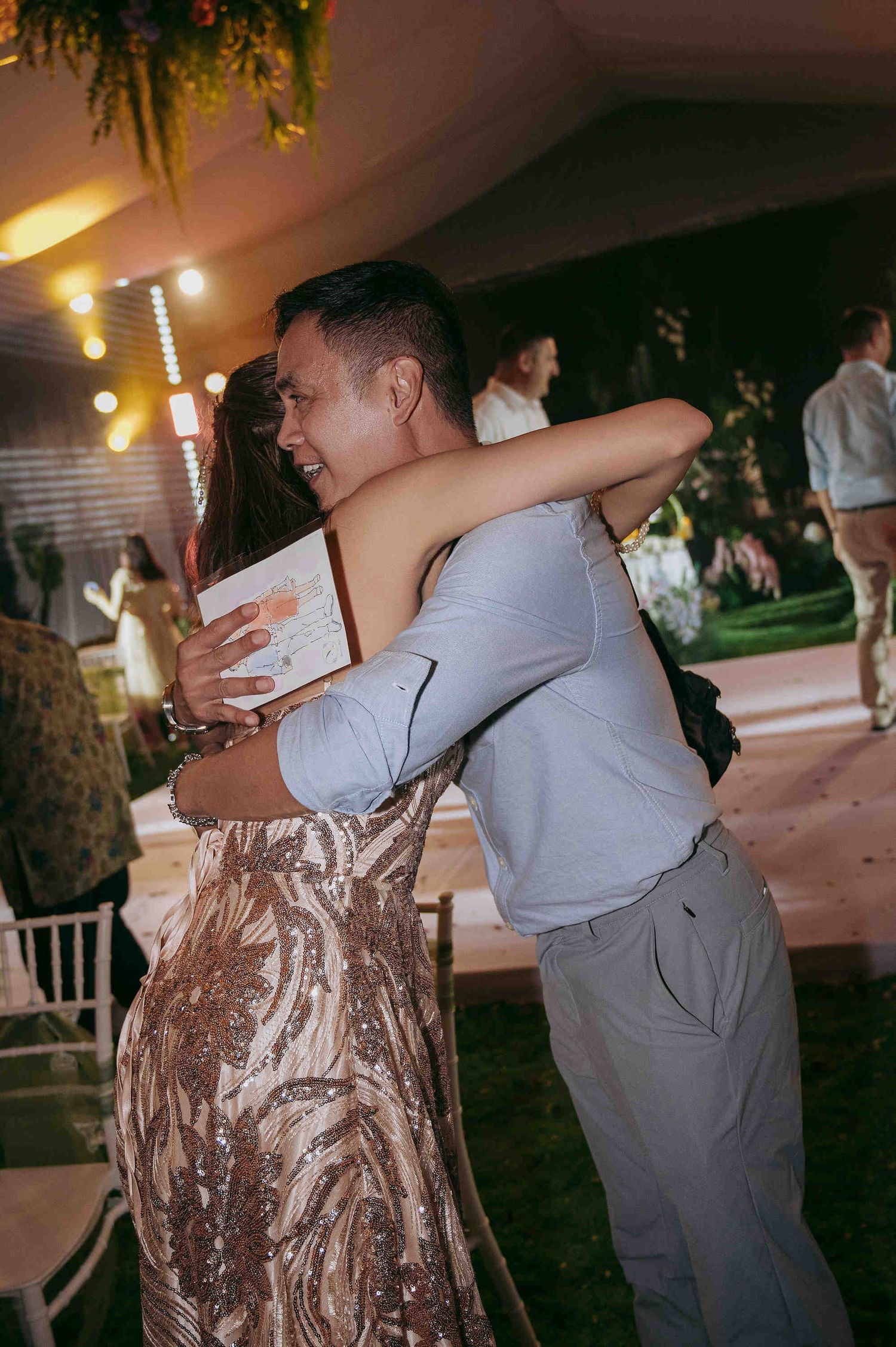 Two guests share a warm hug at an elegant evening wedding reception, woman wearing a gold sequin gown.