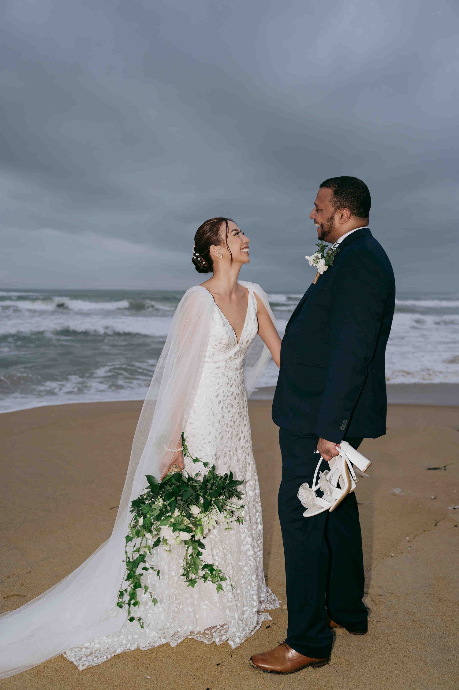 Bride and groom share a joyful moment on a sandy beach, with ocean waves and cloudy skies behind them.