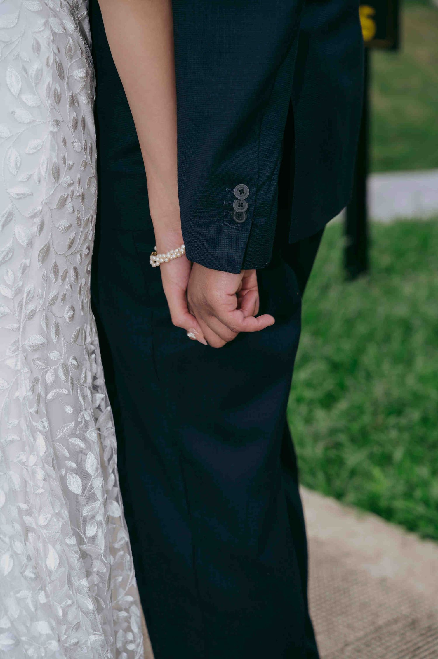 Close-up of a bride and groom holding hands, bride wearing a pearl bracelet and lace wedding gown.