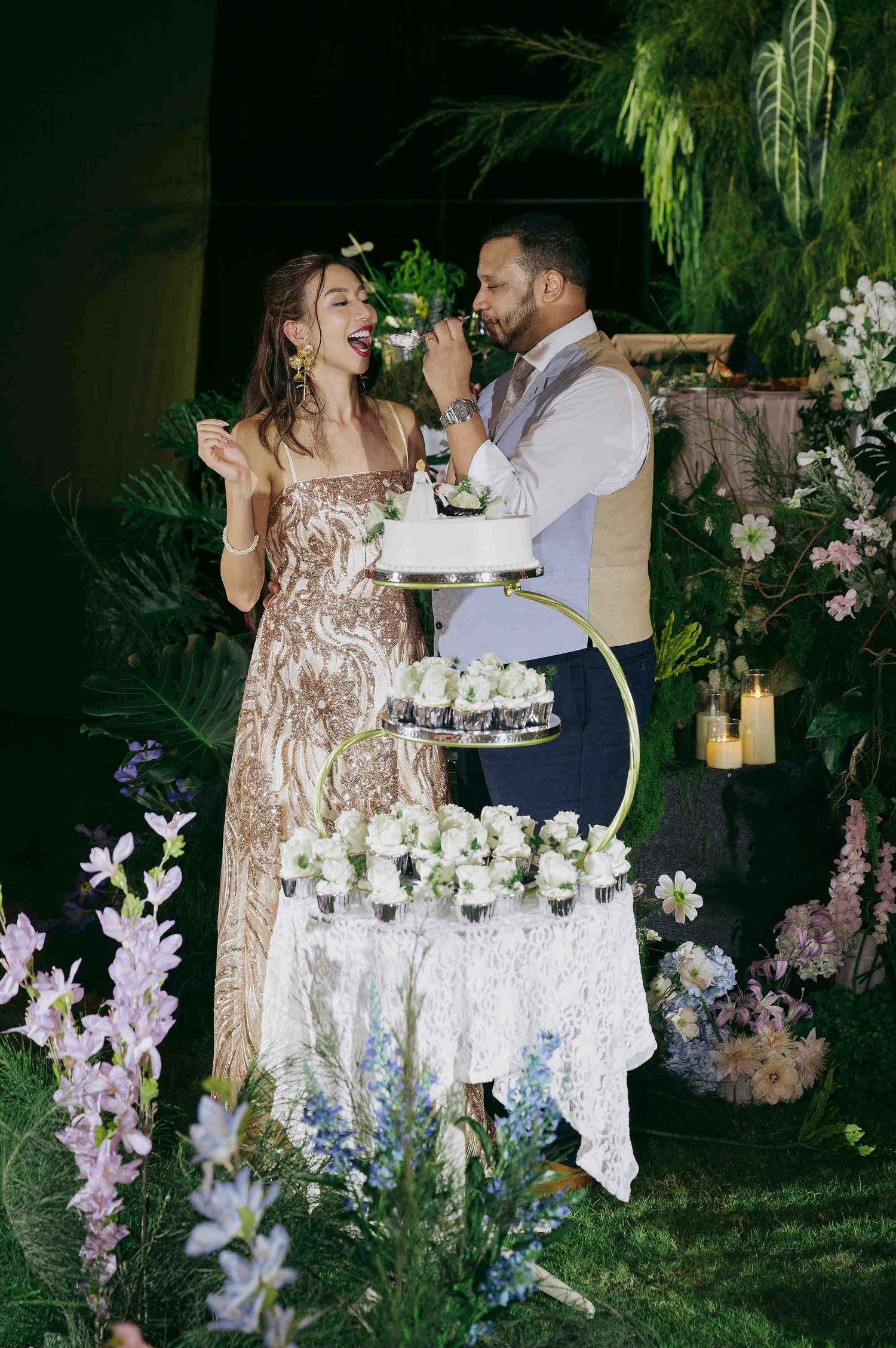 Couple joyfully cutting a floral wedding cake outdoors at night, surrounded by lush greenery and colorful flowers.