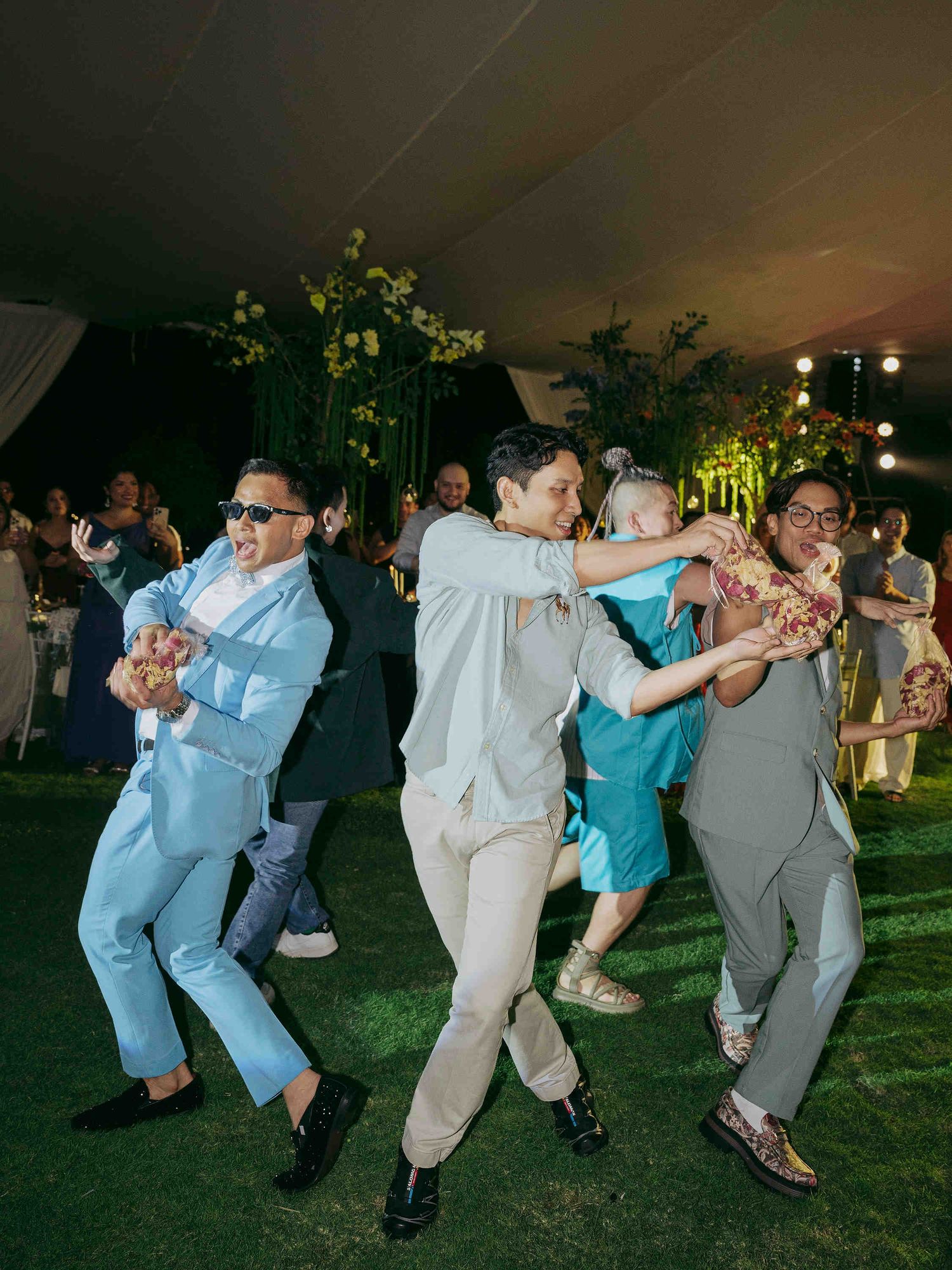Groomsmen dancing energetically at an outdoor wedding reception under a tent with floral decorations at night.