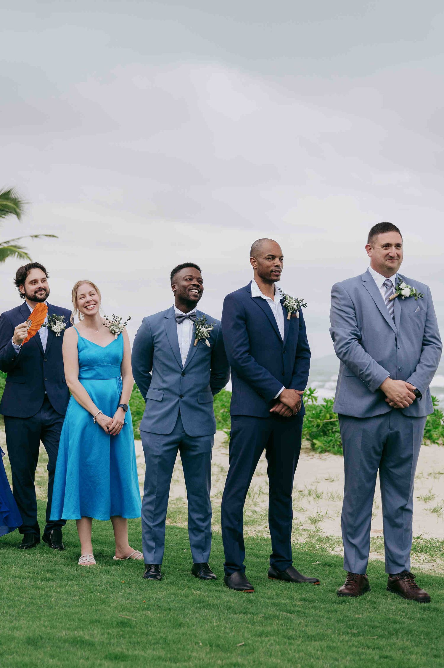 Wedding party standing outdoors near beach, groomsmen in navy and grey suits with a bridesmaid in blue dress.