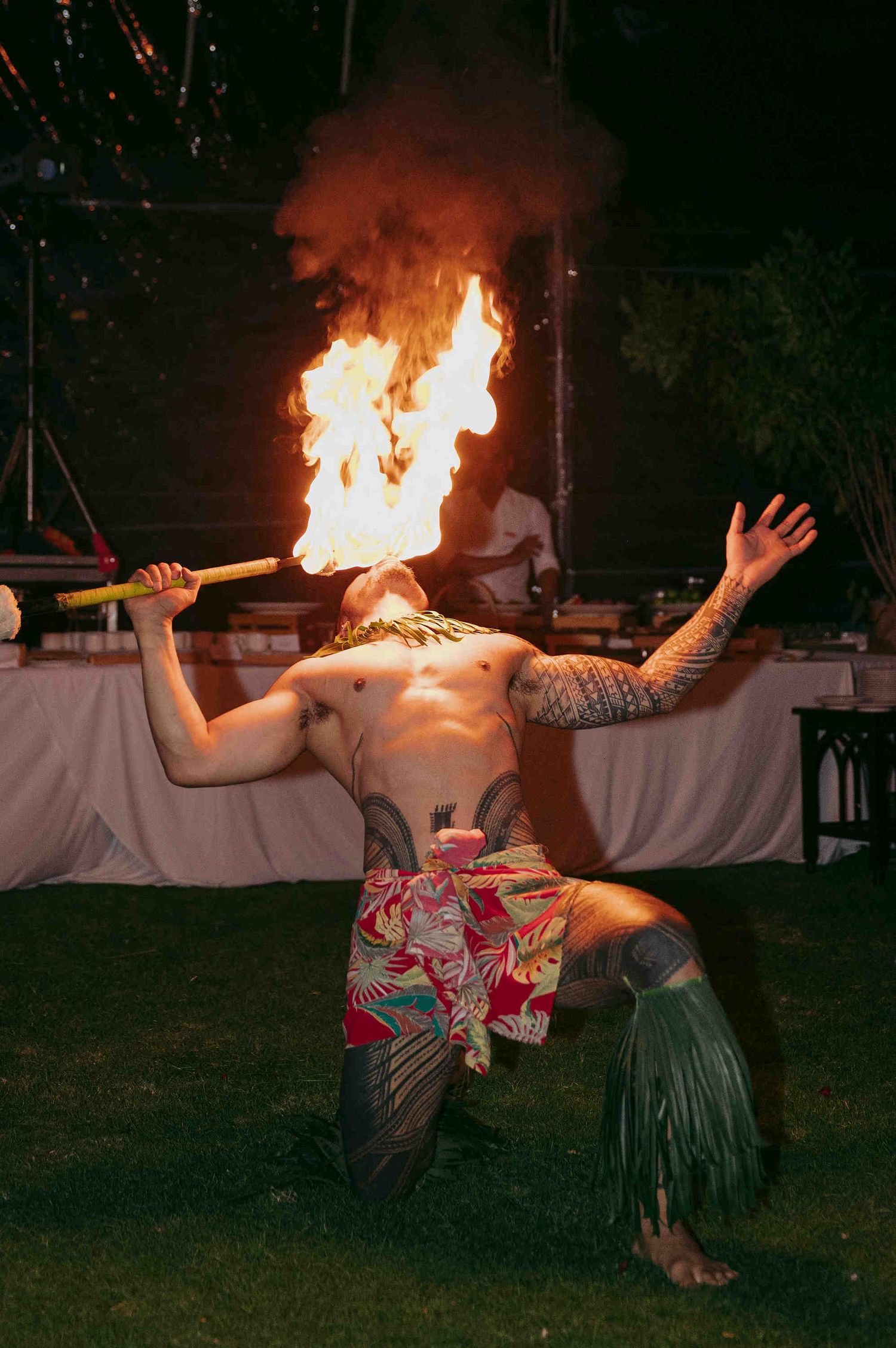 Tattooed fire dancer performs at outdoor luau event, breathing flames while kneeling on grass in traditional Hawaiian attire.