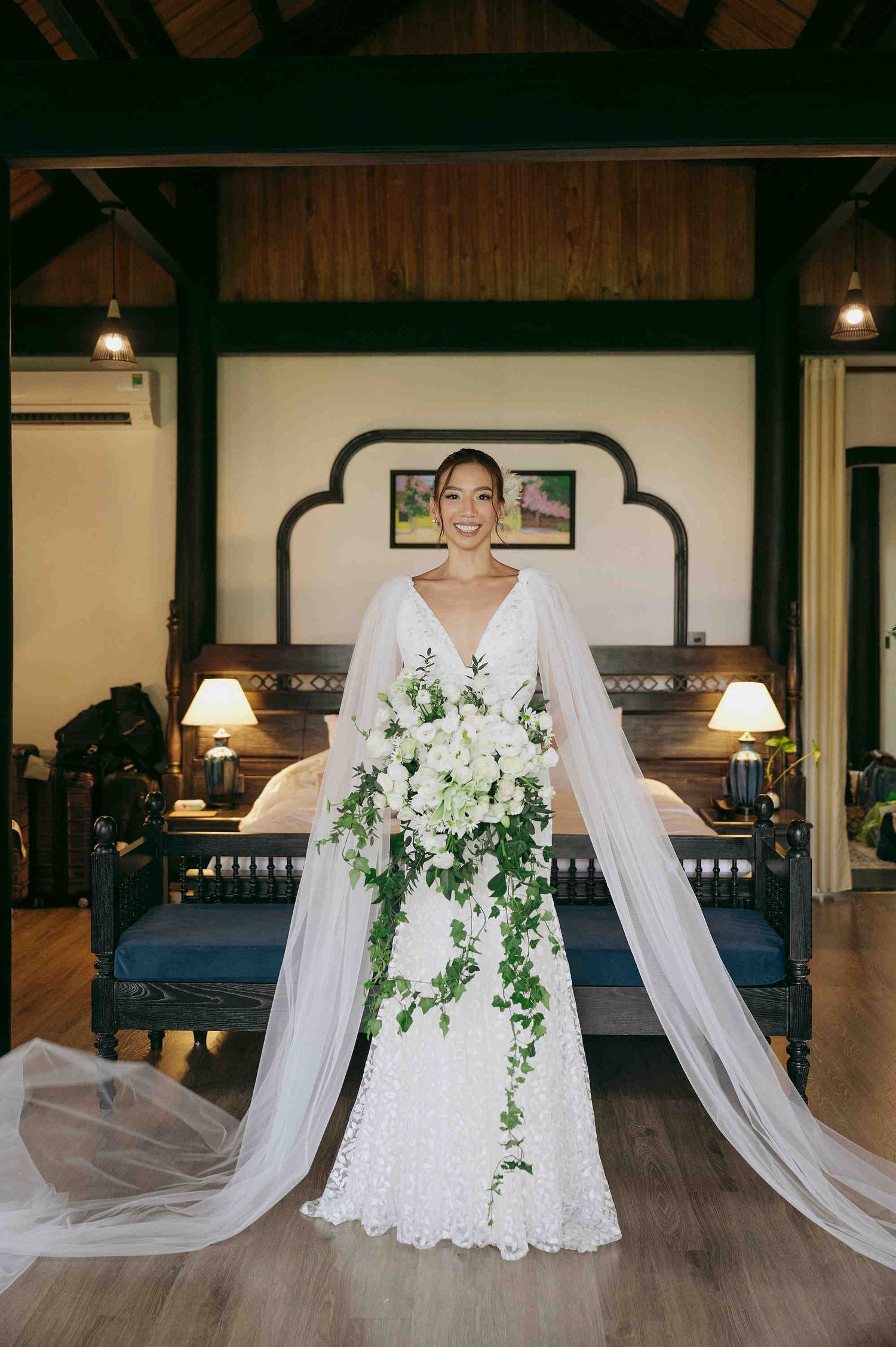 Smiling bride in white lace gown with cape veil holds cascading greenery bouquet in a luxurious hotel suite.