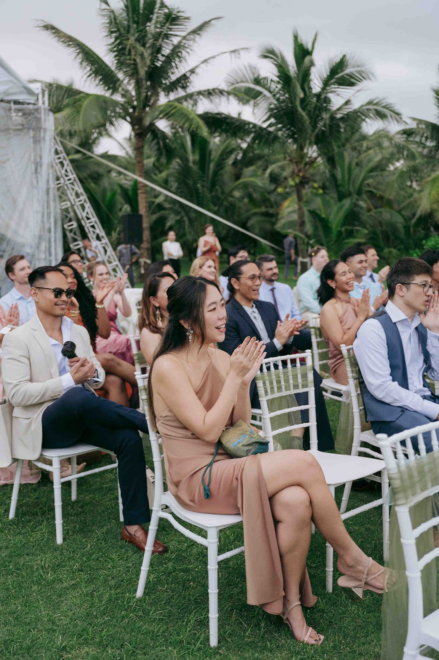 Guests clapping and smiling at an outdoor wedding ceremony on a tropical lawn with palm trees.