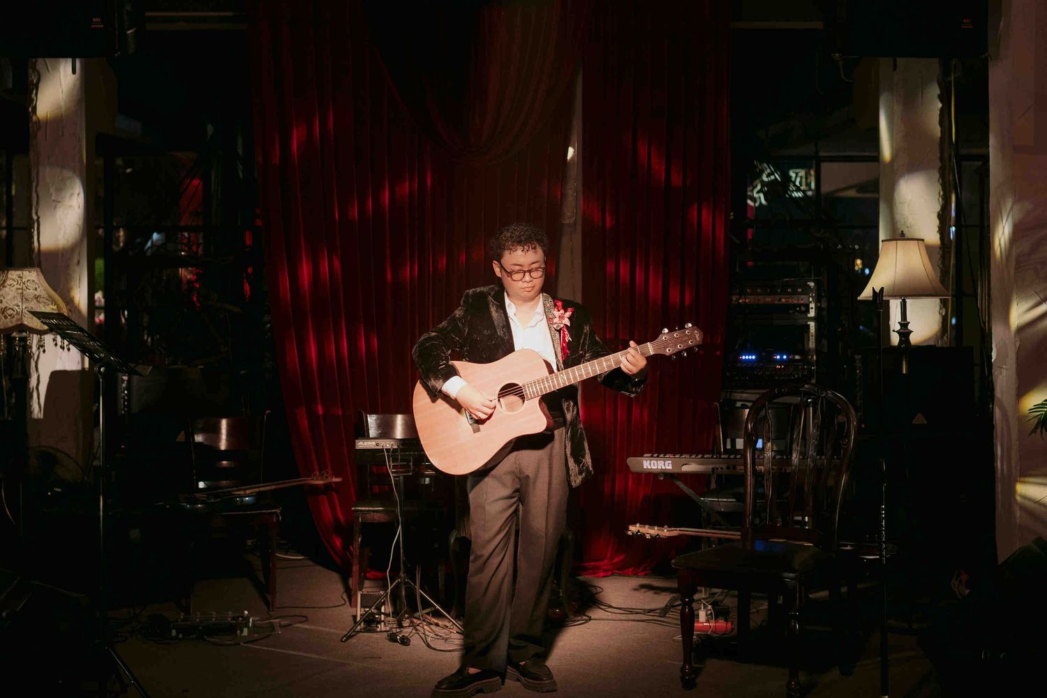 A musician plays acoustic guitar on a dimly lit stage with red curtains backdrop.