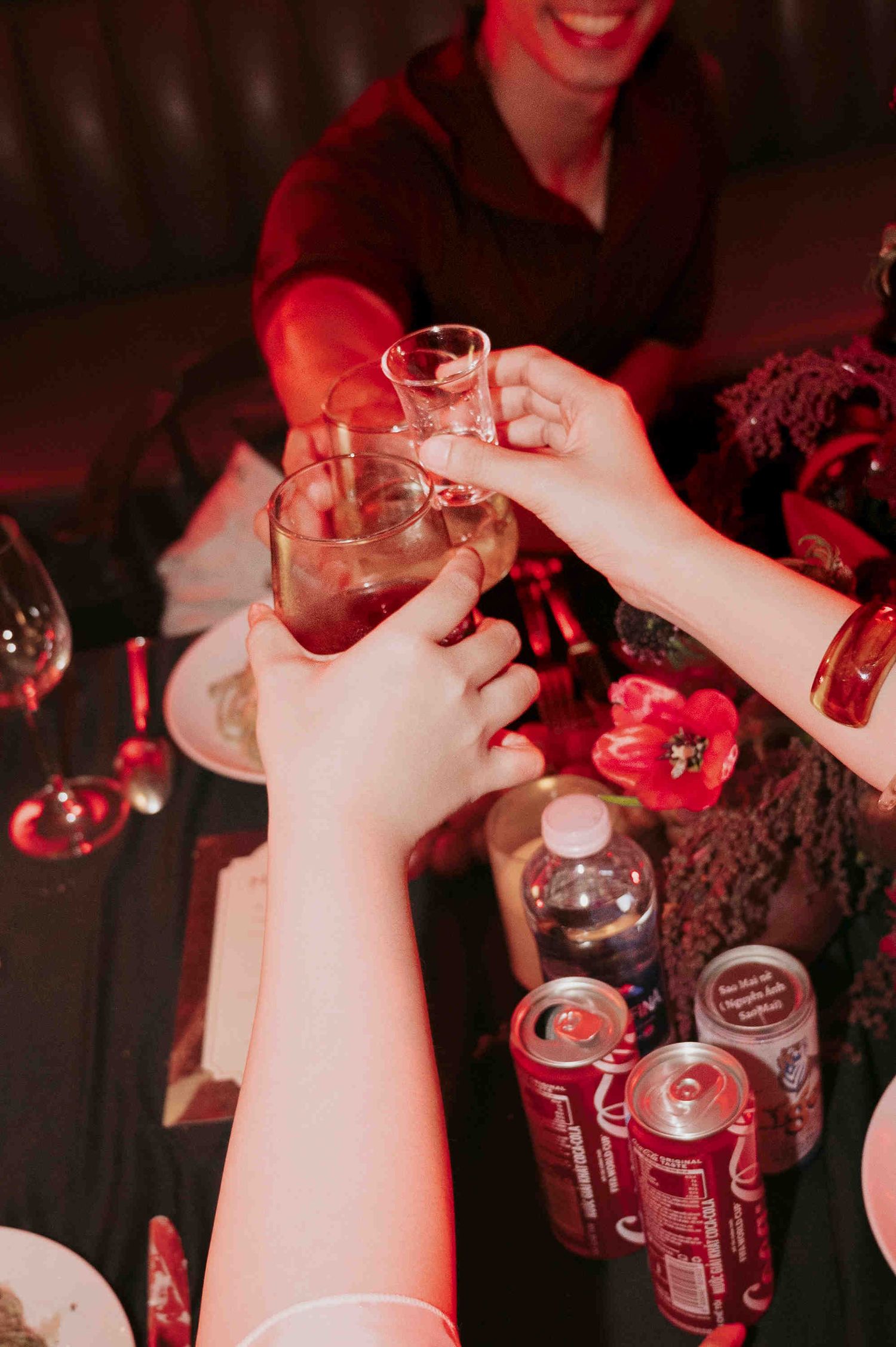 Friends toasting with cocktails and shot glasses at a dimly lit bar table with red ambient lighting.