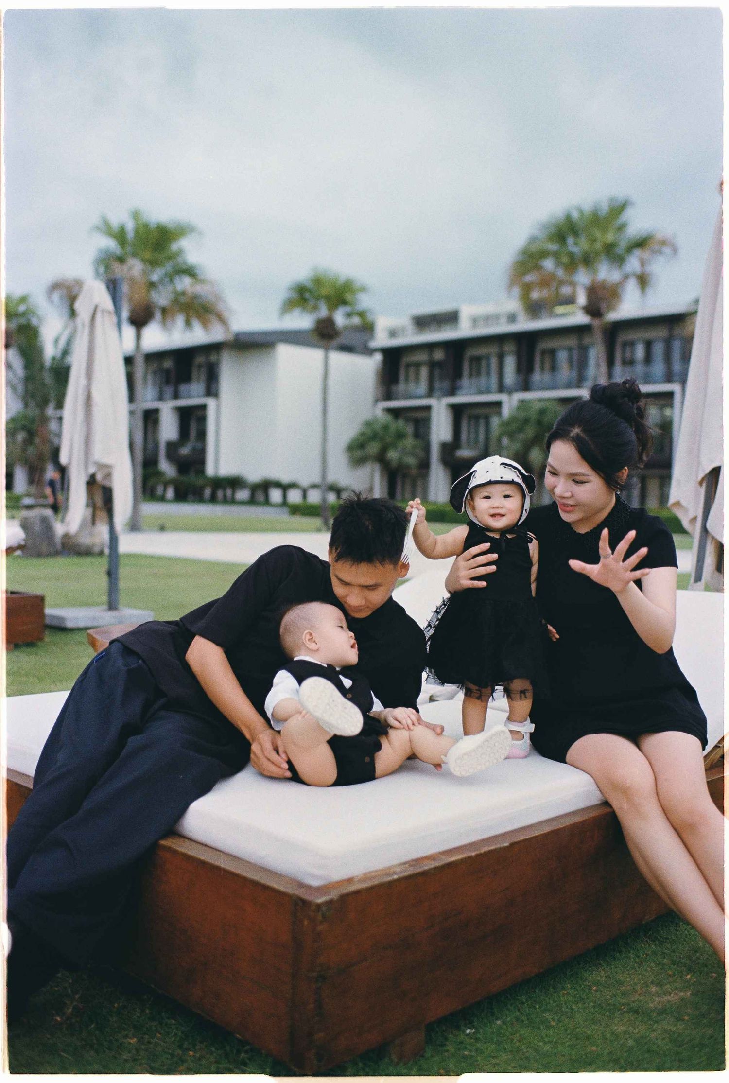 Da Nang family: family wearing black clothing plays together on an outdoor concrete bench surrounded by palm trees, by AnhVuWedding.