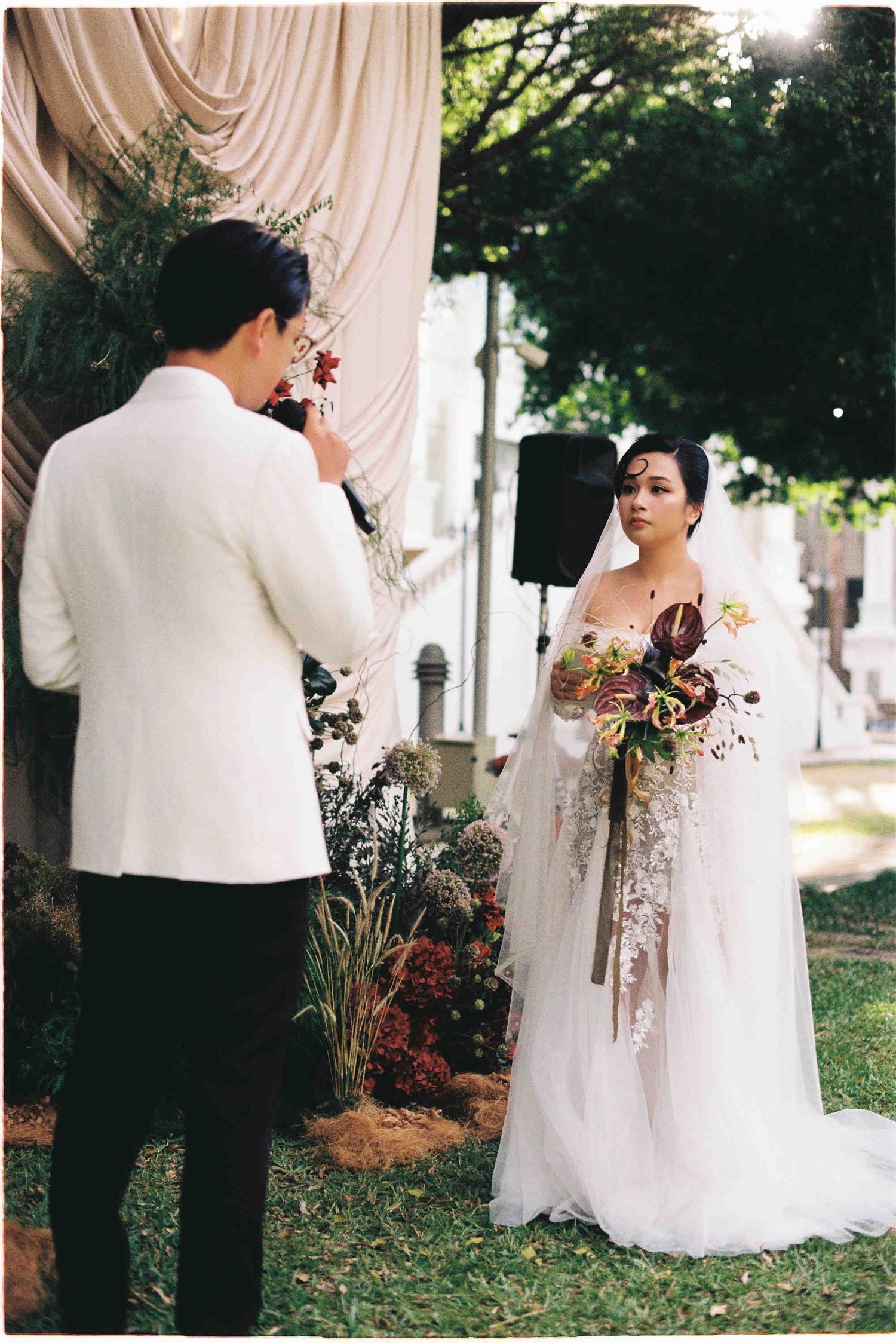 Bride in lace wedding gown holding bouquet exchanges vows with groom in white barong at outdoor garden ceremony.