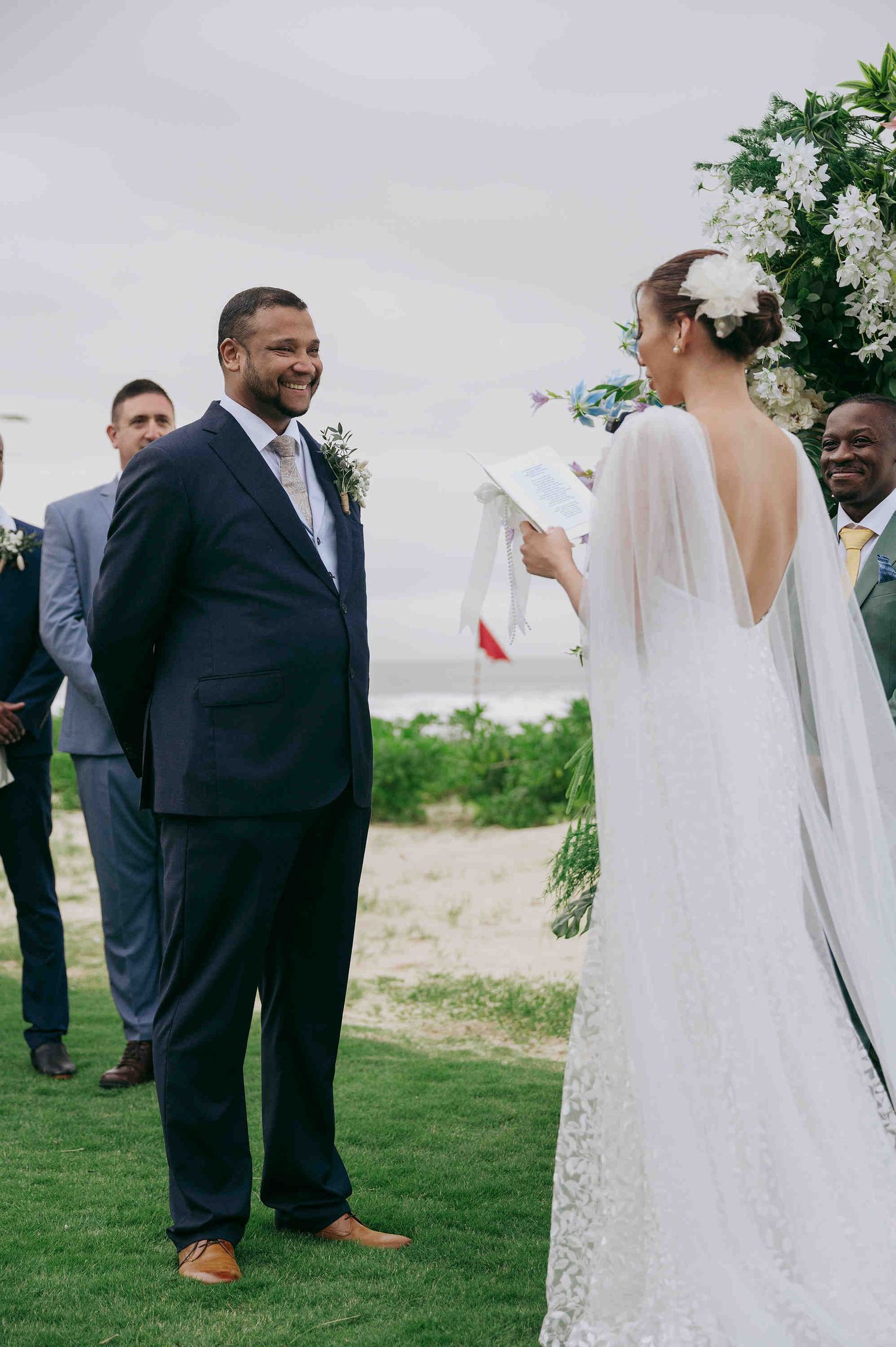 Groom in navy suit smiling at bride in backless wedding gown during outdoor beach ceremony.