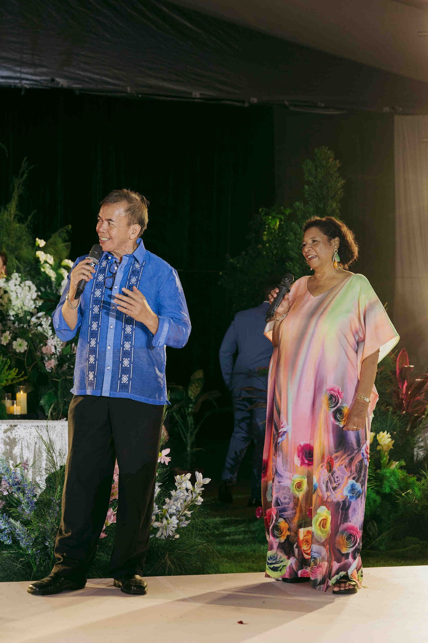 Man in blue barong and woman in floral kaftan speak at microphones during an elegant evening event.