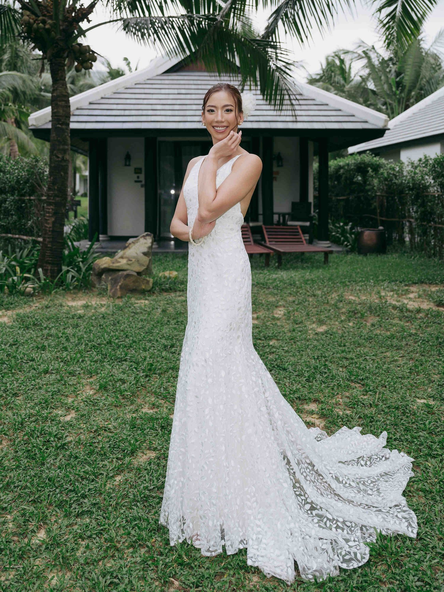 Bride in a fitted white lace wedding gown with a train poses on green lawn near tropical bungalow.