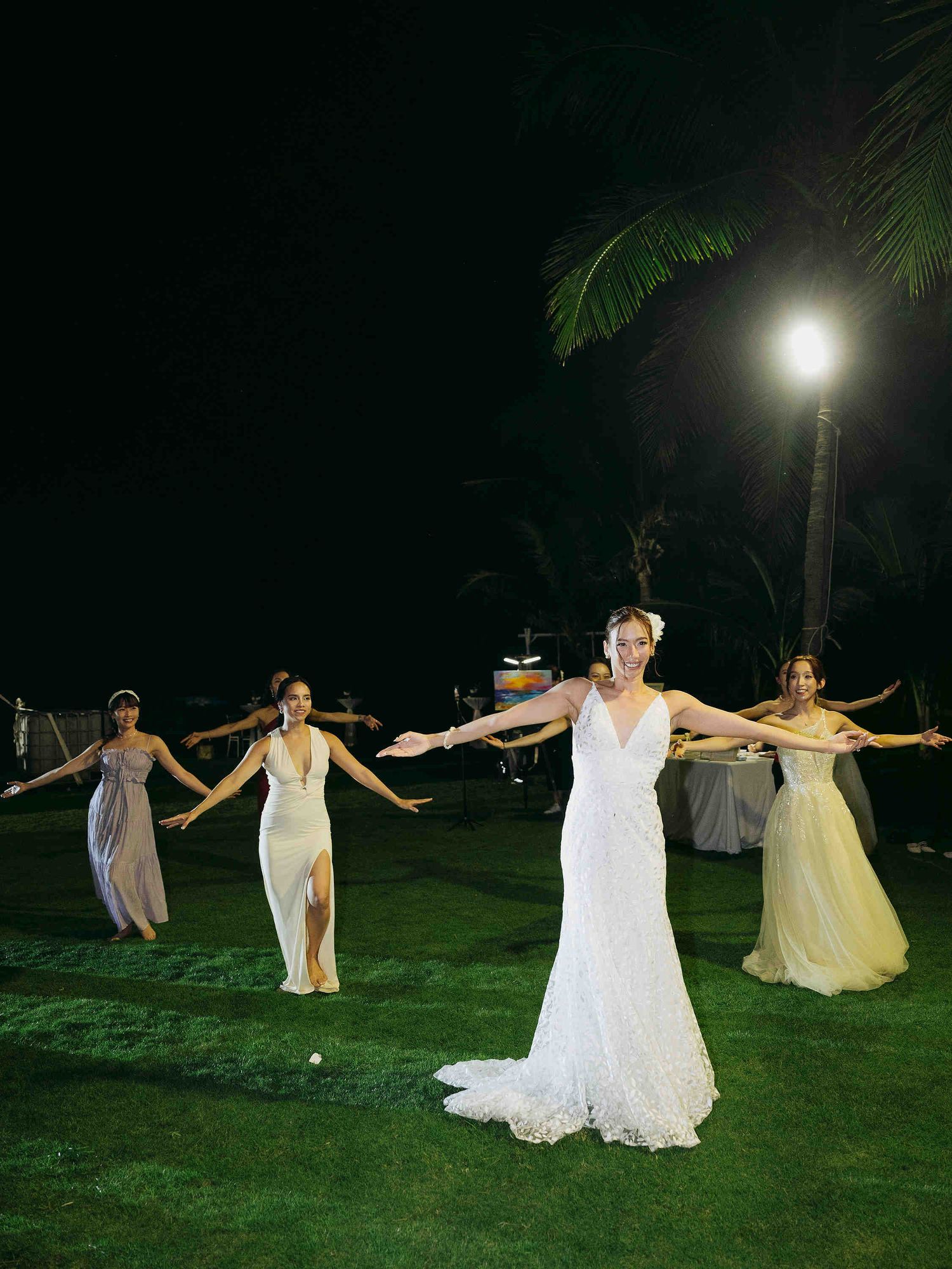 Bride in white lace gown poses with bridesmaids in elegant dresses at an outdoor nighttime wedding reception.
